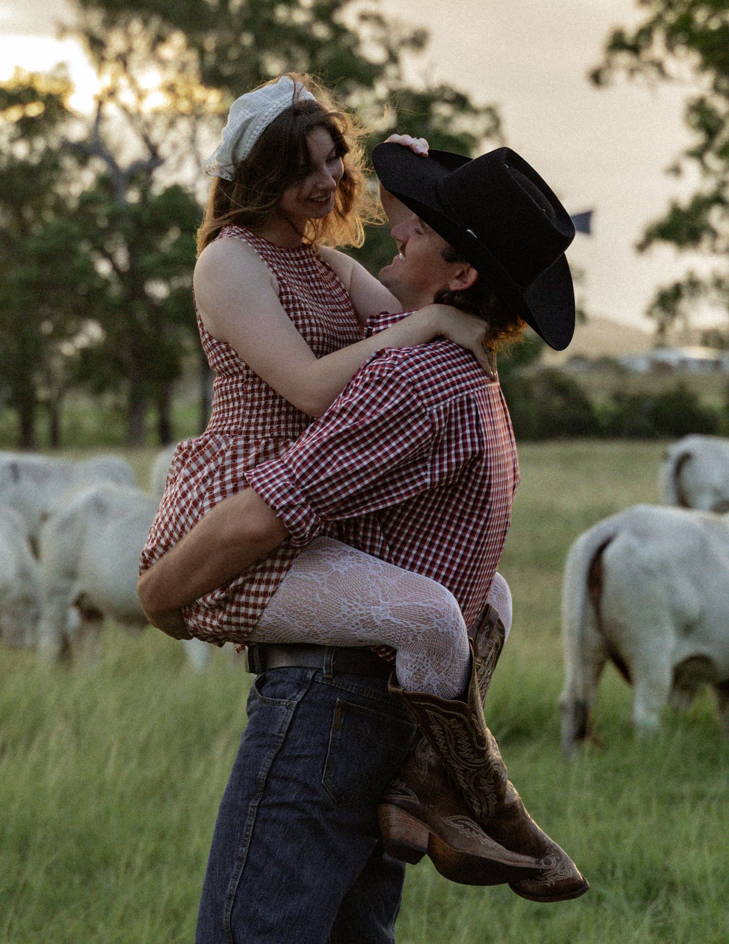 Cowboy in a black hat lifts a woman in a gingham dress and lace tights in a golden-lit pasture with white cattle behind them.