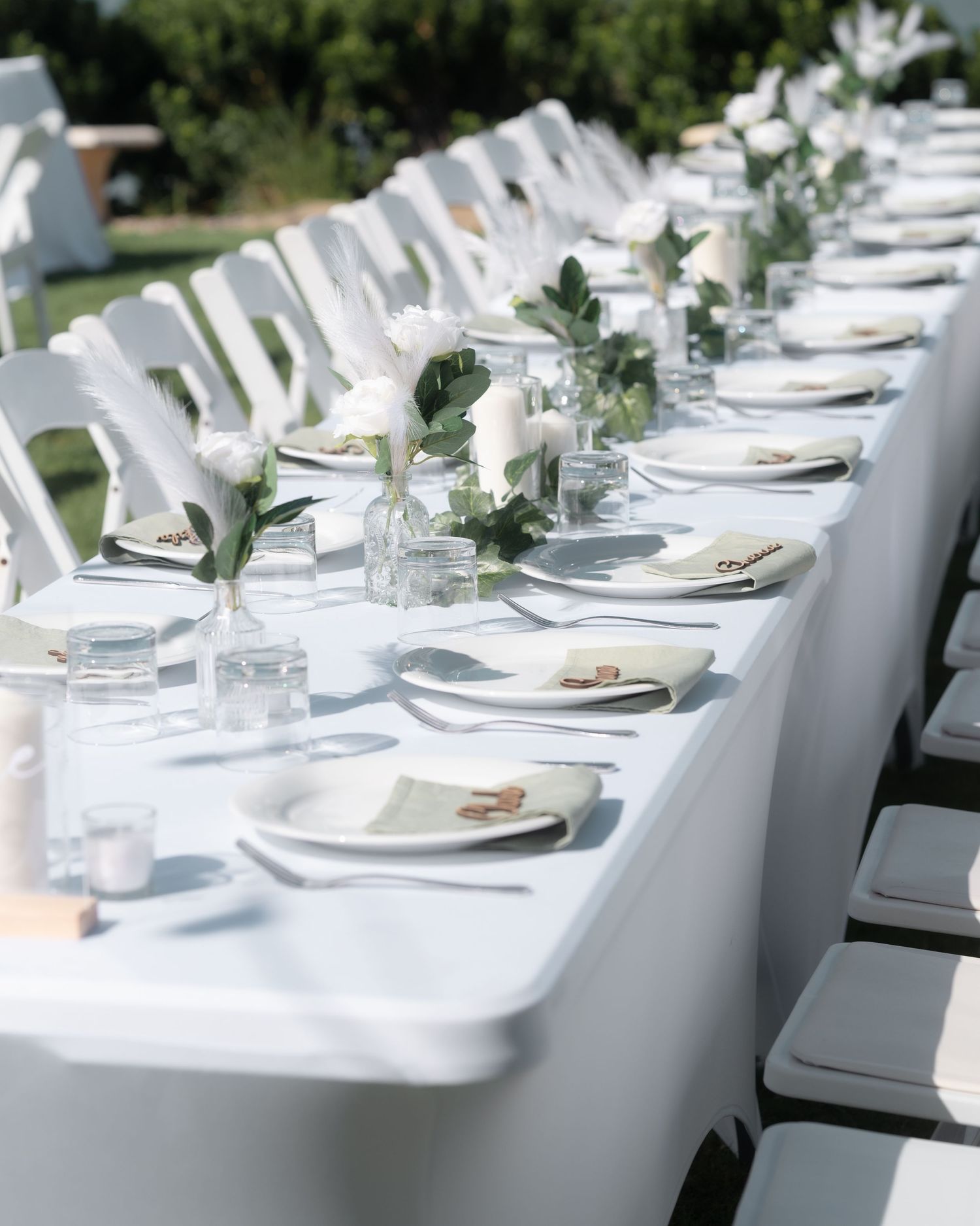 Long white wedding reception table set with plates, green napkins, glassware and small white floral arrangements in vases.