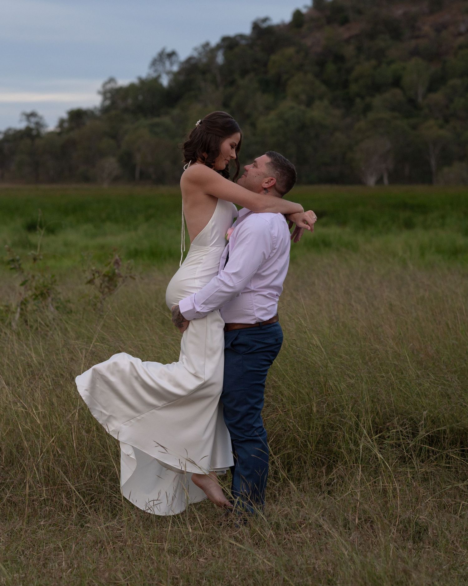 Groom lifts his bride in a white wedding dress off the ground in an embrace in a green field with hills behind.
