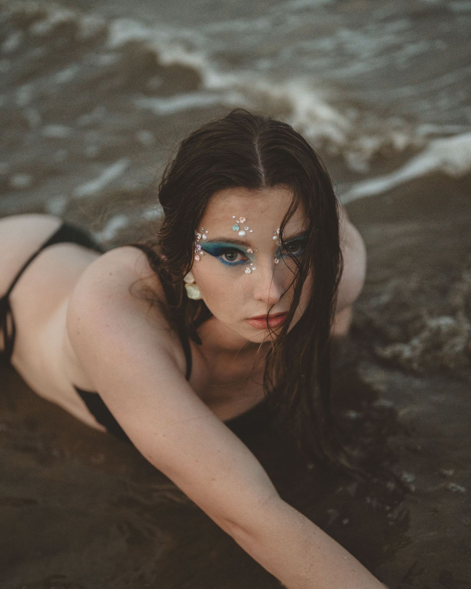 Woman with dark wet hair and sparkling face gems lies on wet sand as ocean waves roll in behind her.
