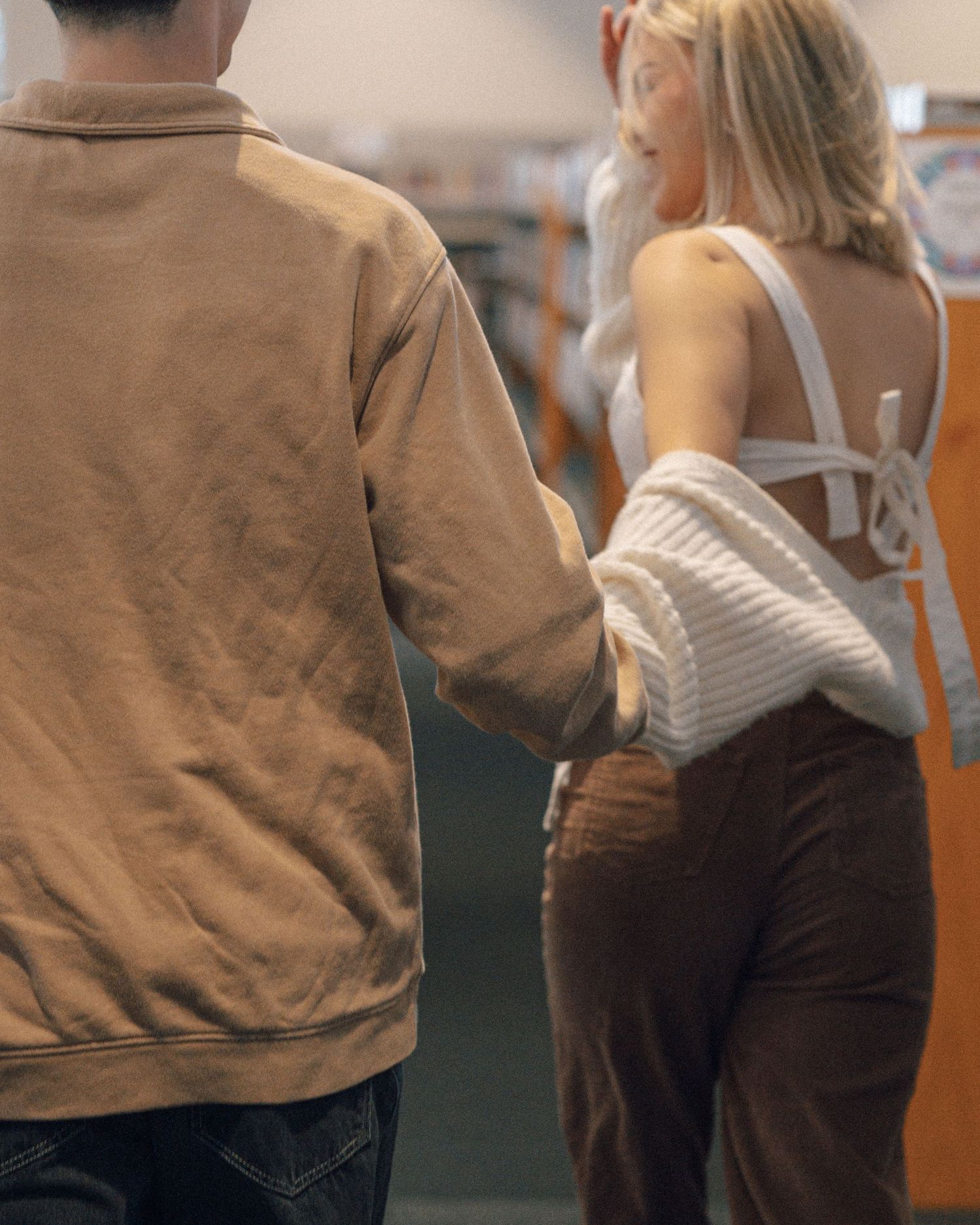A couple holds hands and walks through a library, the woman in a white cardigan and brown corduroy pants.