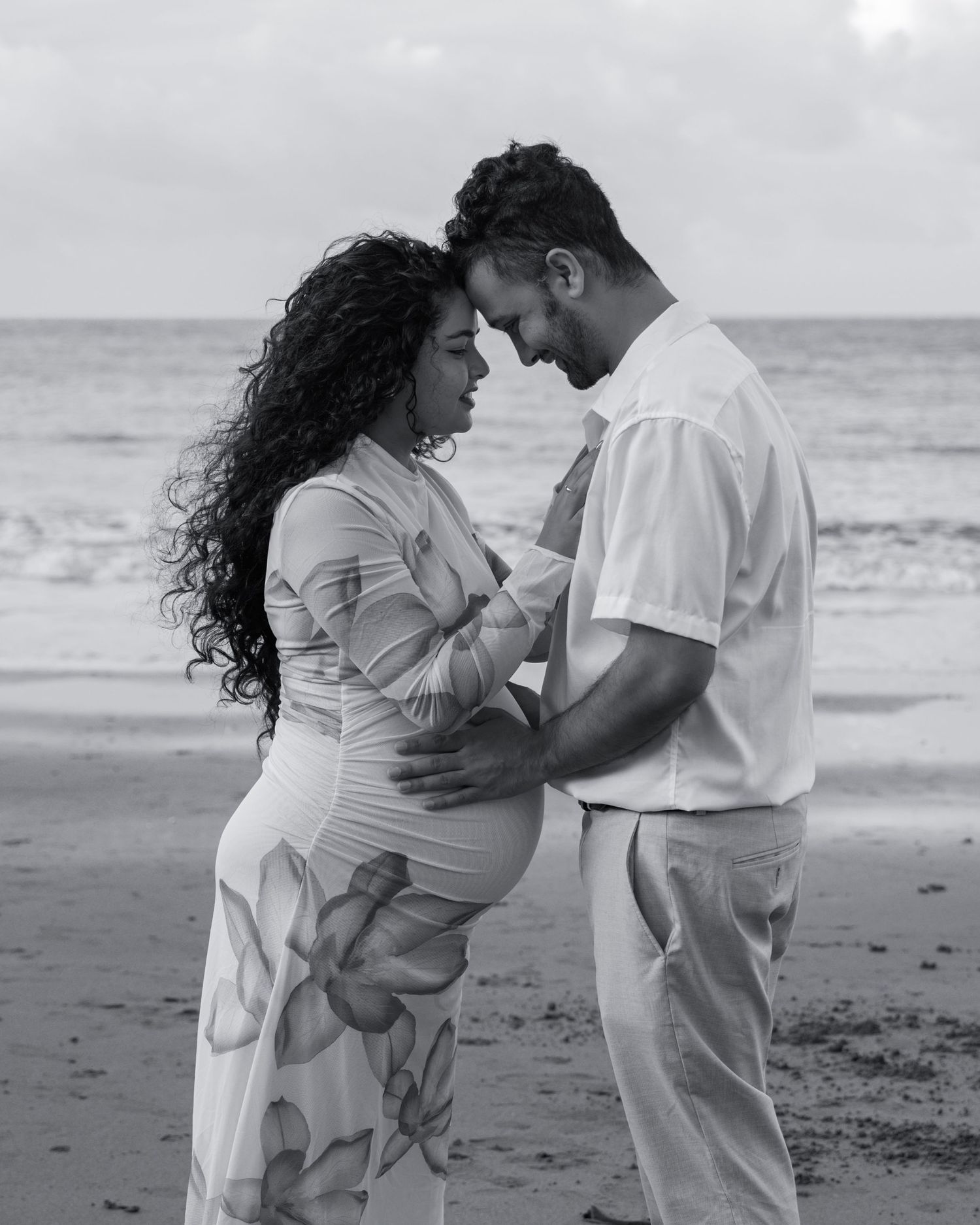Black and white beach maternity photo of an expectant couple touching foreheads as the husband cradles her belly.