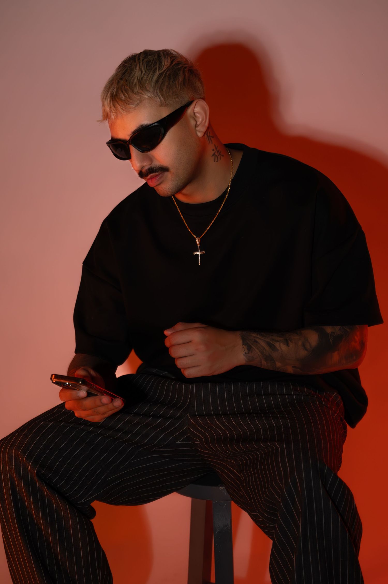 Man in black sunglasses, oversized black tee and pinstriped trousers sits on a stool checking his phone against a warm red-orange backdrop.