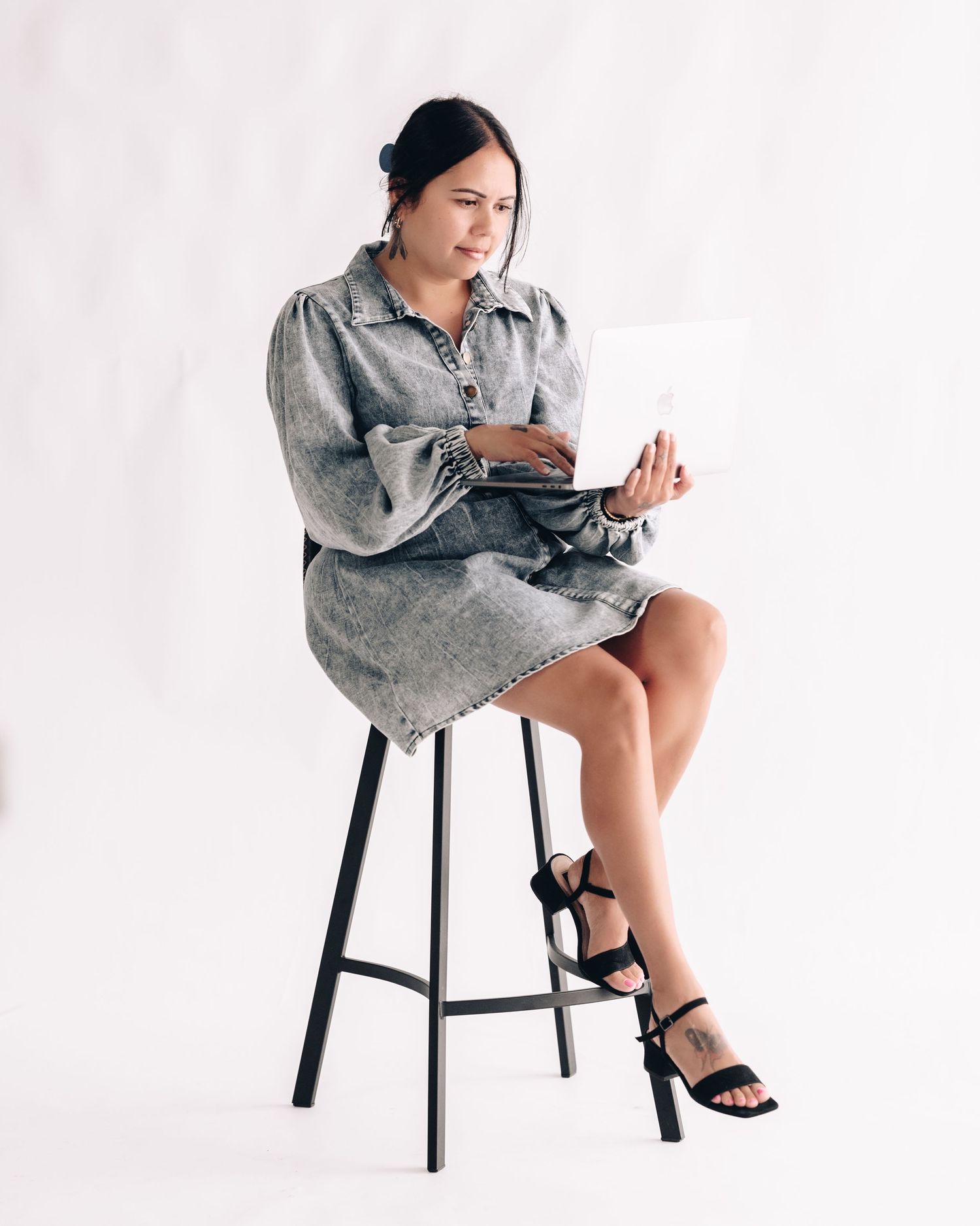 Woman in a gray denim dress and black heeled sandals sits on a stool working on a laptop against a white backdrop.