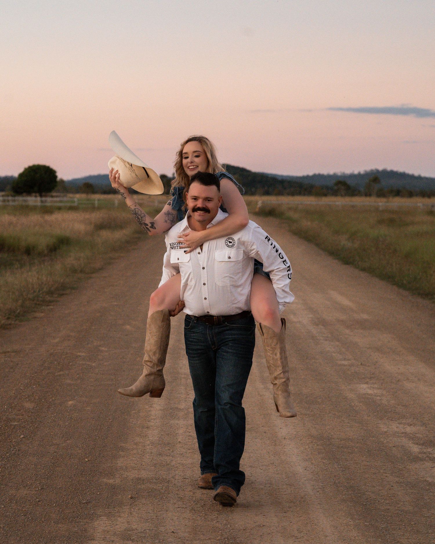 Smiling man in a western shirt carries a laughing woman piggyback along a dirt road at sunset during a country engagement shoot.