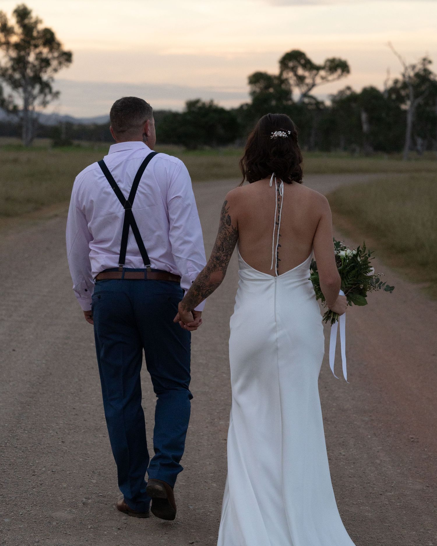 Bride holding a bouquet and groom in suspenders walk hand in hand down a gravel country road at dusk.