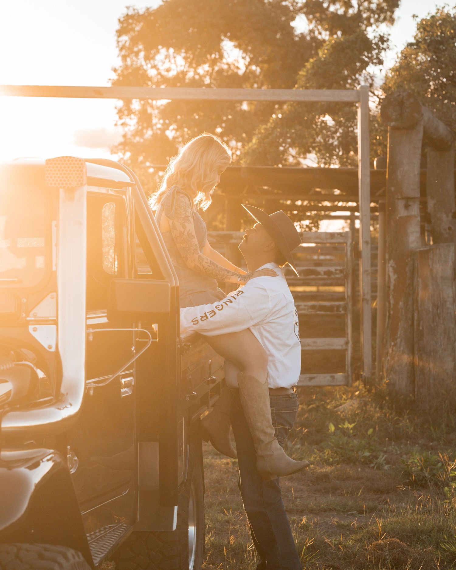 A woman in a denim dress wraps her leg around a cowboy as they embrace by a ranch gate in golden sunset light.