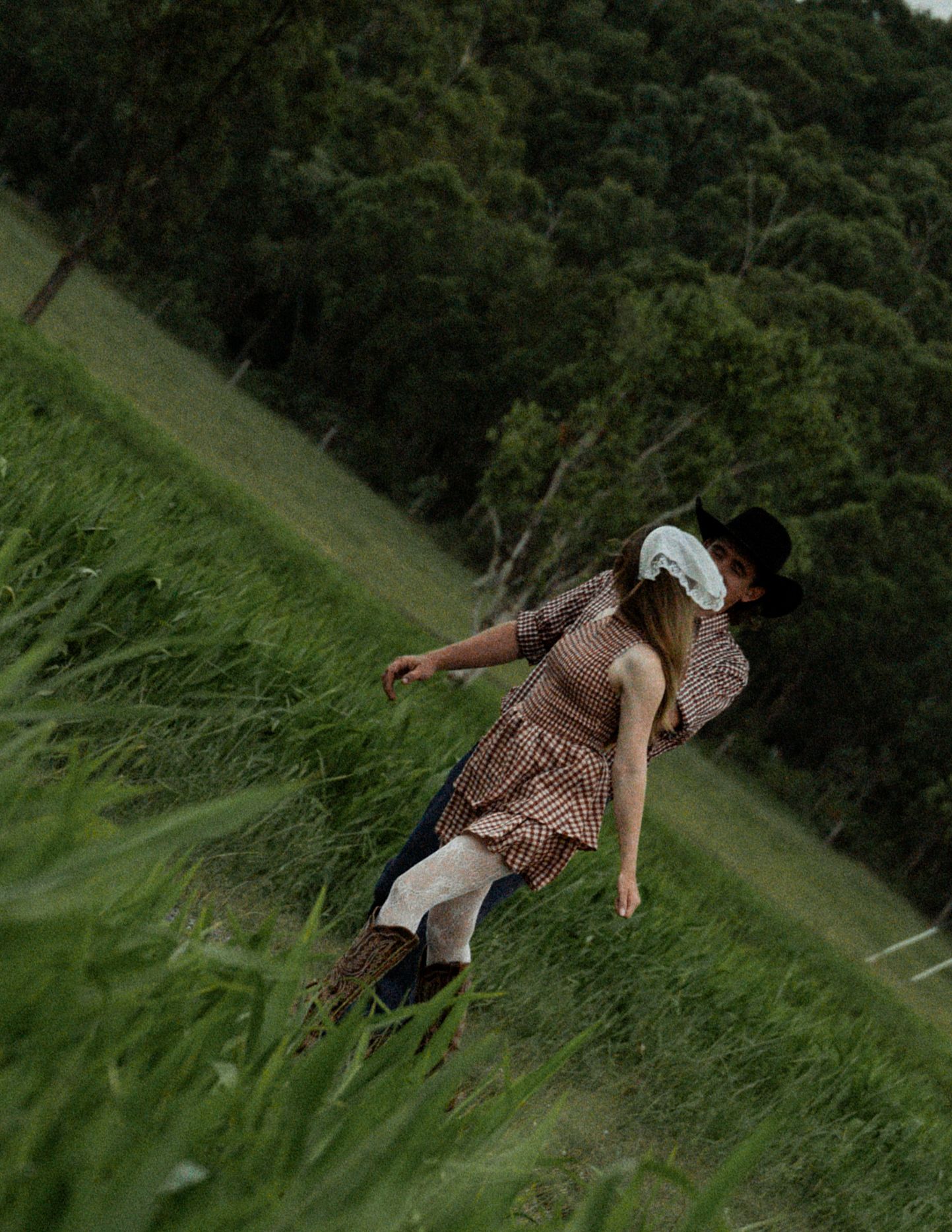 A woman in a gingham dress and white lace head covering walks through tall green grass beside a man in a black cowboy hat.