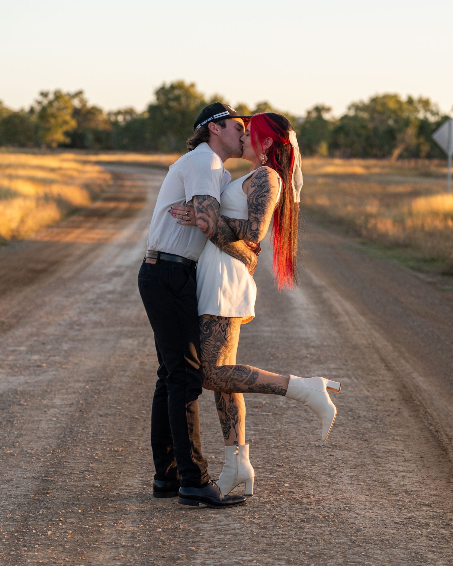 Tattooed couple kissing on a country dirt road at golden hour, the woman with red hair lifting her leg in white boots.