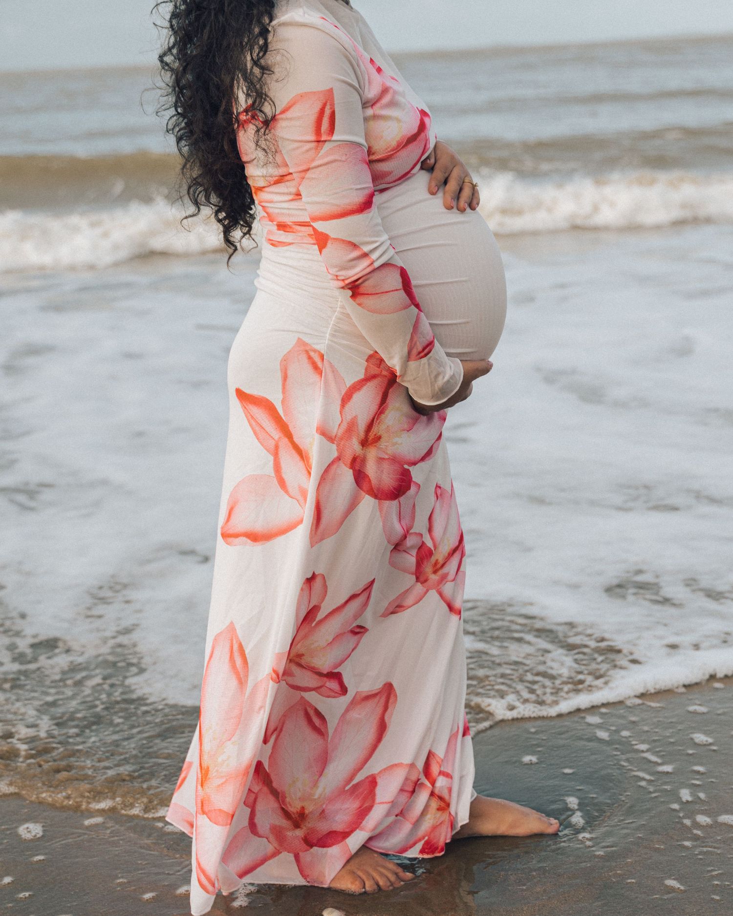 Barefoot pregnant woman in a long coral floral dress stands at the water's edge as gentle waves wash over the sandy beach.