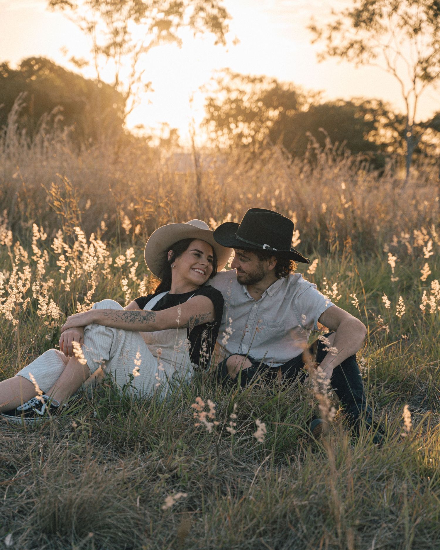 Smiling couple in cowboy hats sit close together in a golden sunset field of tall grass and wildflowers.