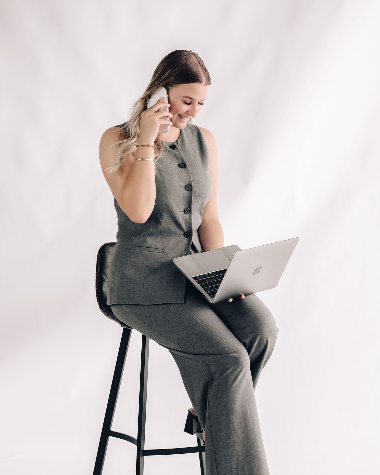 Woman in a gray vest and matching trousers sits on a black stool, smiling while on a phone call with a laptop on her lap.