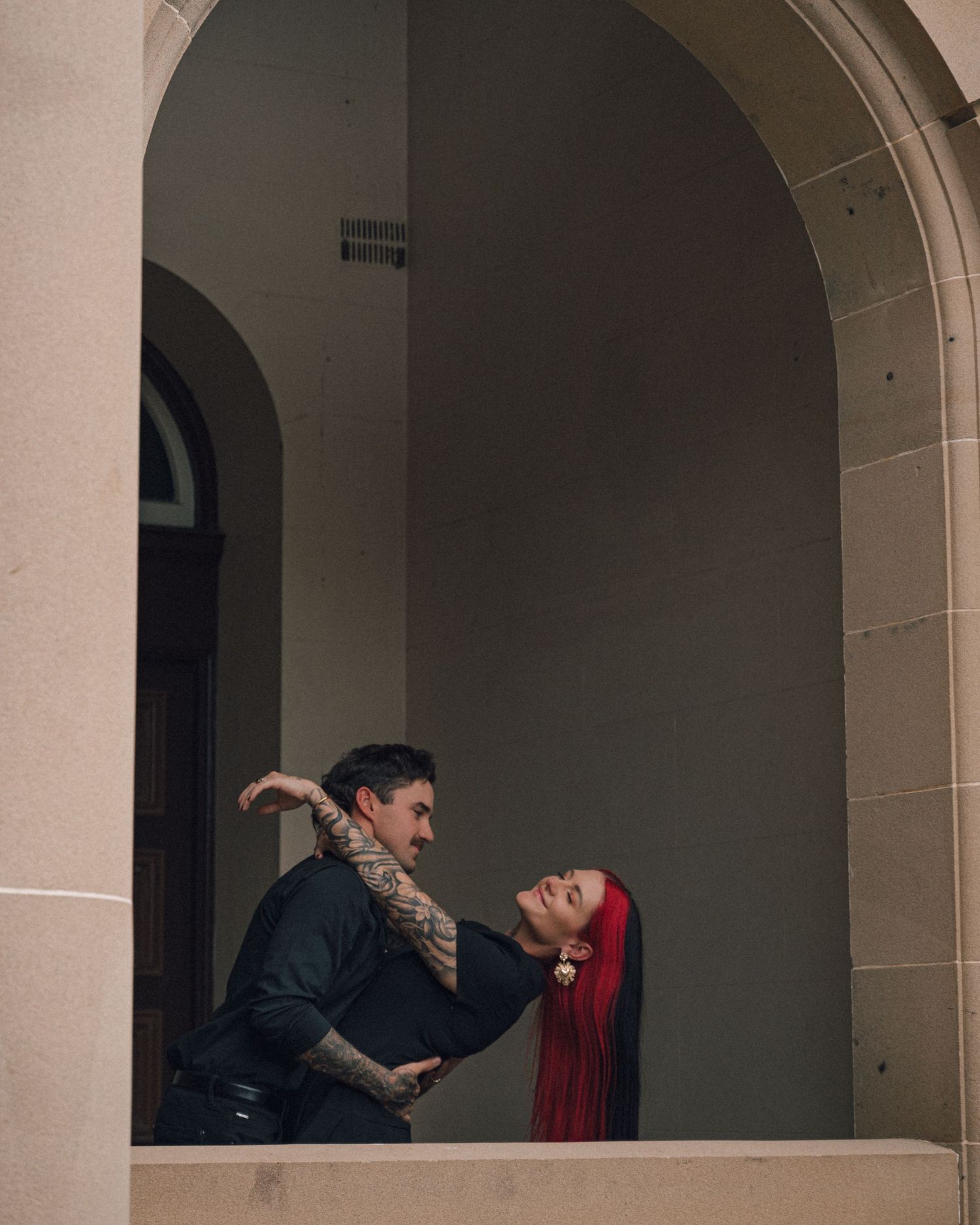 Couple with tattoos and red hair share a dramatic dip pose inside a sandstone archway.