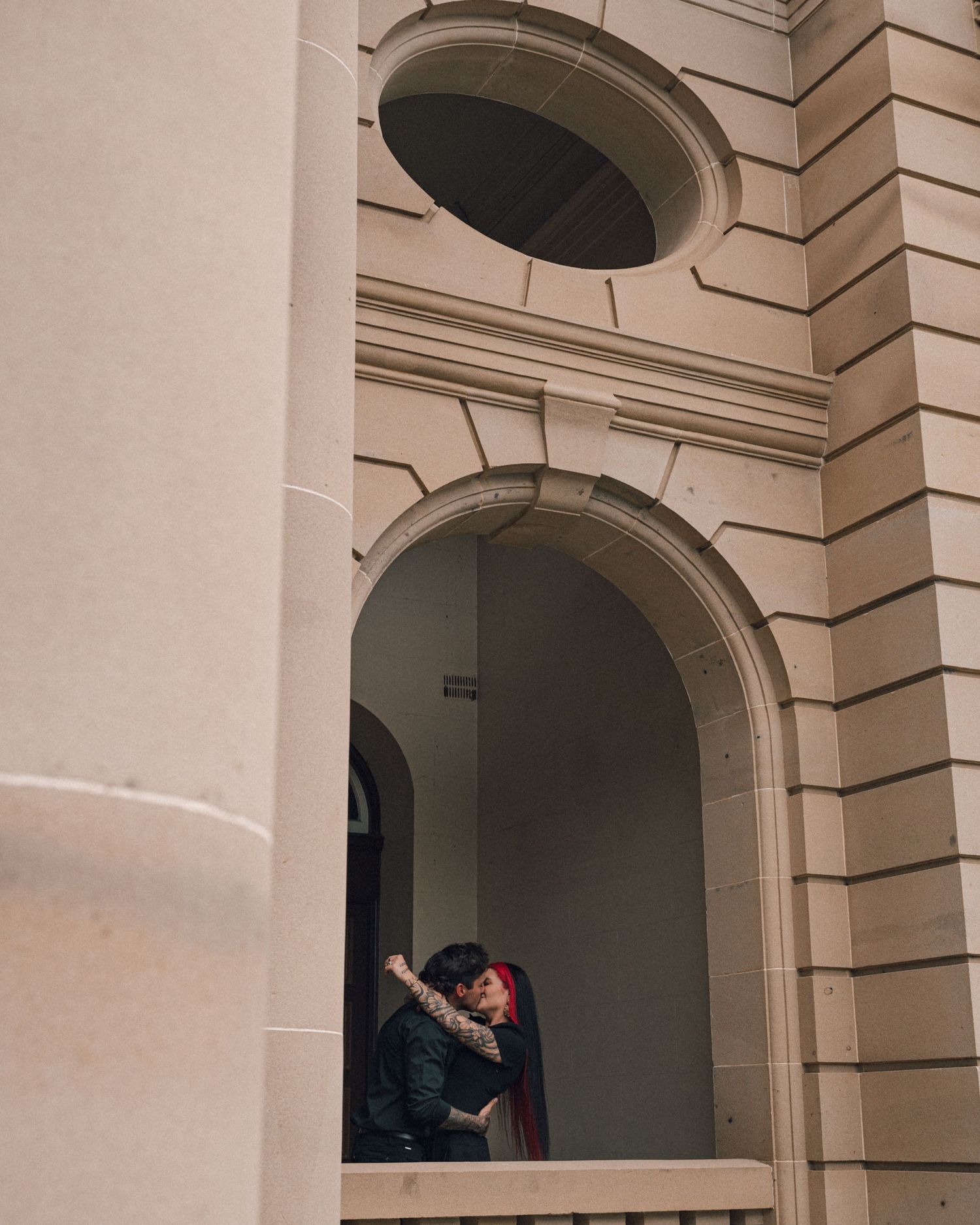 A tattooed couple embraces in a stone archway beneath a round window on a classic sandstone building with elegant architectural details.