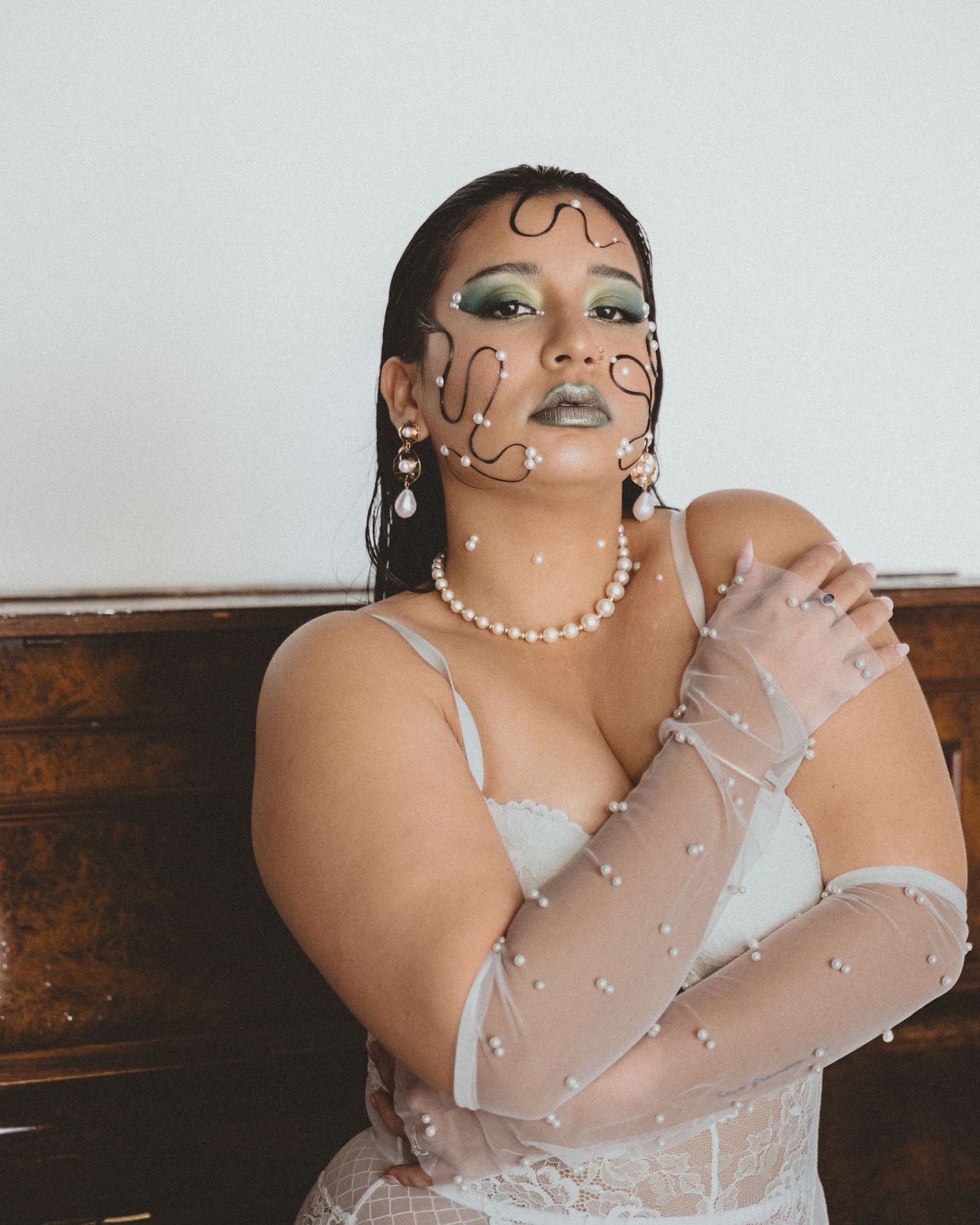 Woman with sculptural black wire face jewelry, green eyeshadow, pearl necklace and sheer pearl-studded gloves poses beside a wooden piano.
