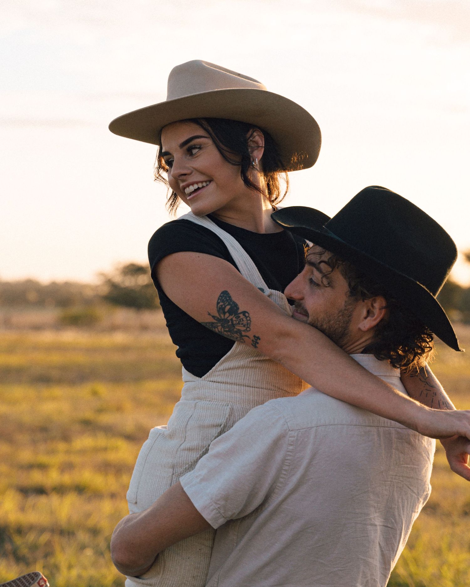 Smiling woman with a tattooed arm and tan hat embraces a bearded man in a black cowboy hat at sunset.