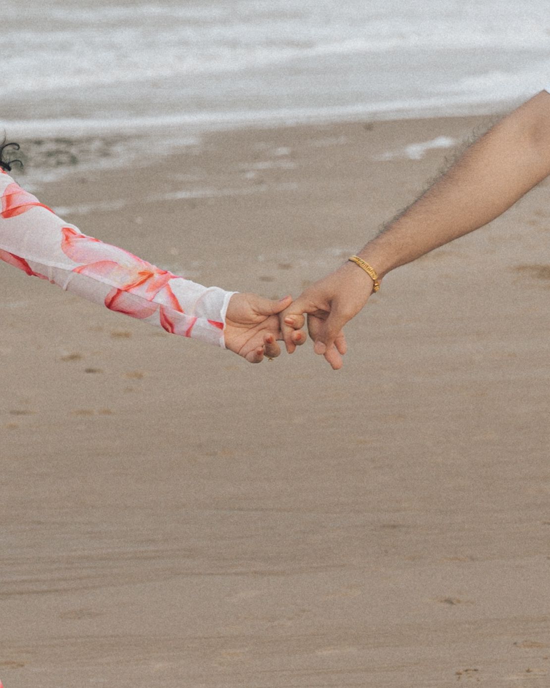 Close-up of a couple holding hands on a sandy beach, her sleeve in pink floral print and his wrist wearing a gold bracelet.