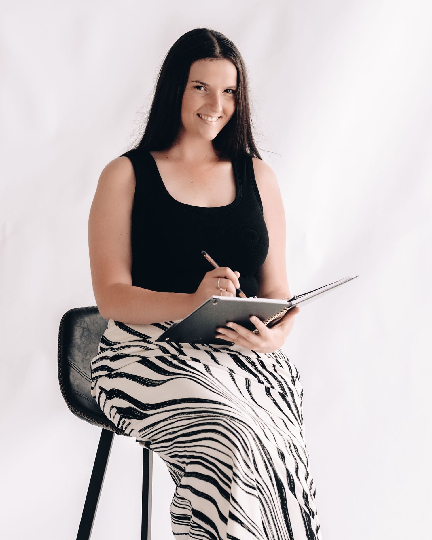 Smiling woman in a black top and zebra-print skirt sits on a stool, holding a pen and writing in an open notebook.