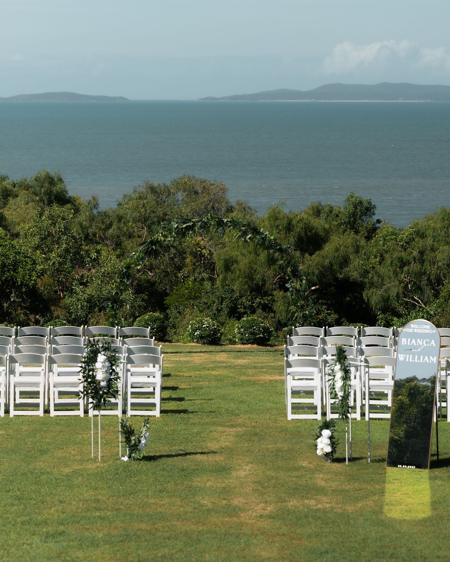 Outdoor wedding ceremony setup with white folding chairs on a lawn overlooking the sea, with a welcome sign naming Bianca and William.