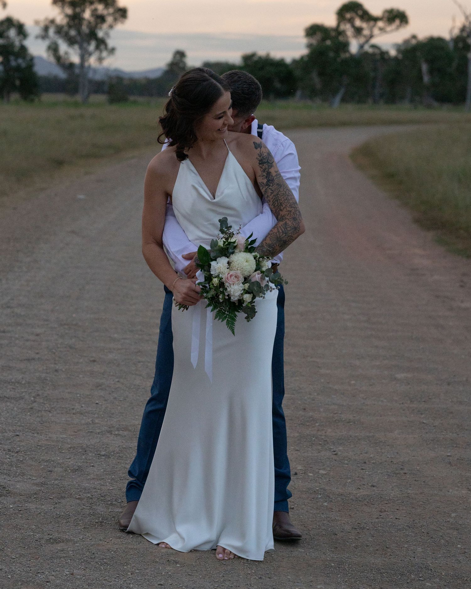 Groom hugs his bride from behind as she holds a white floral bouquet on a country road at dusk.