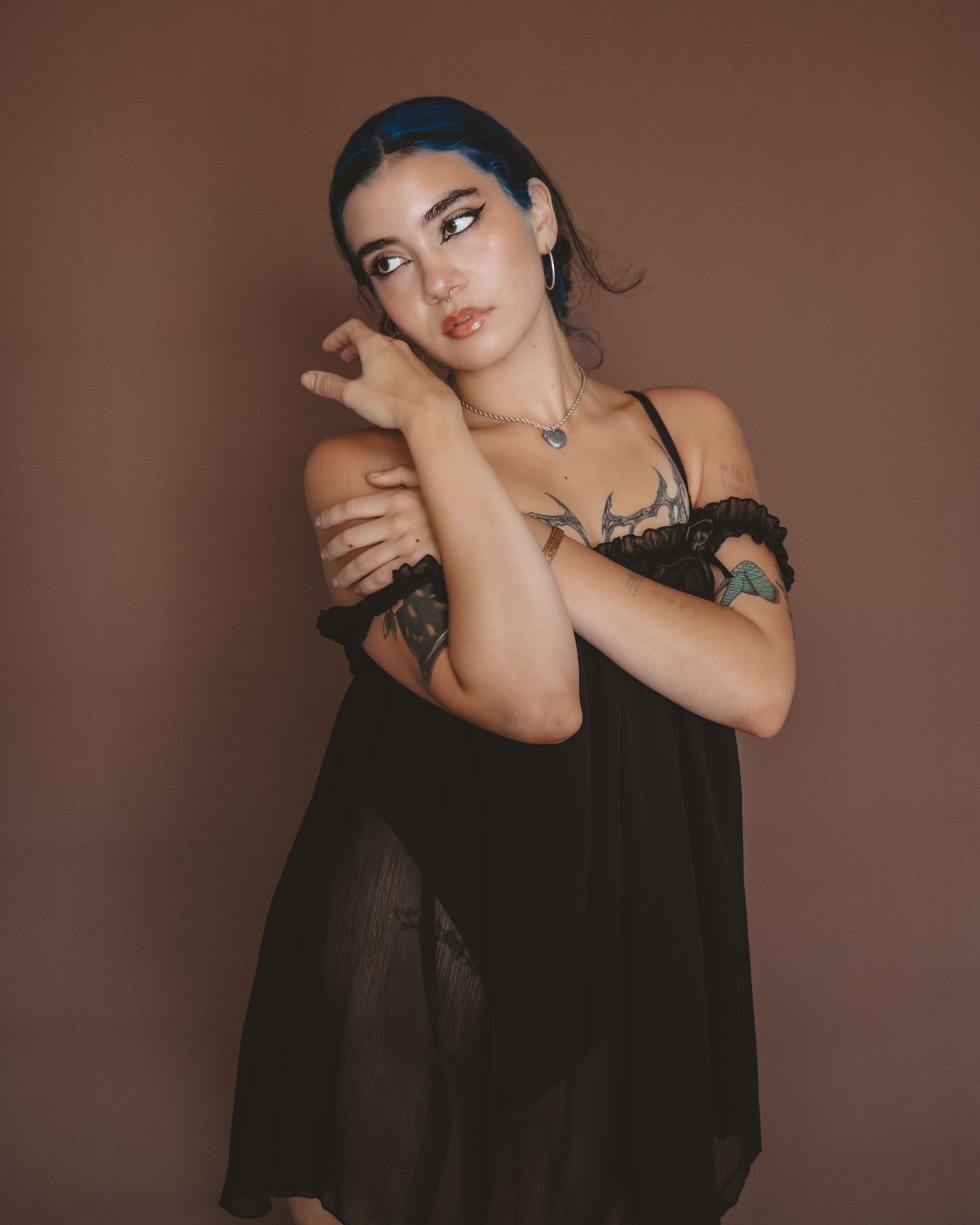 Tattooed woman with blue-streaked hair and graphic eye makeup poses in a sheer black ruffled top against a brown backdrop.