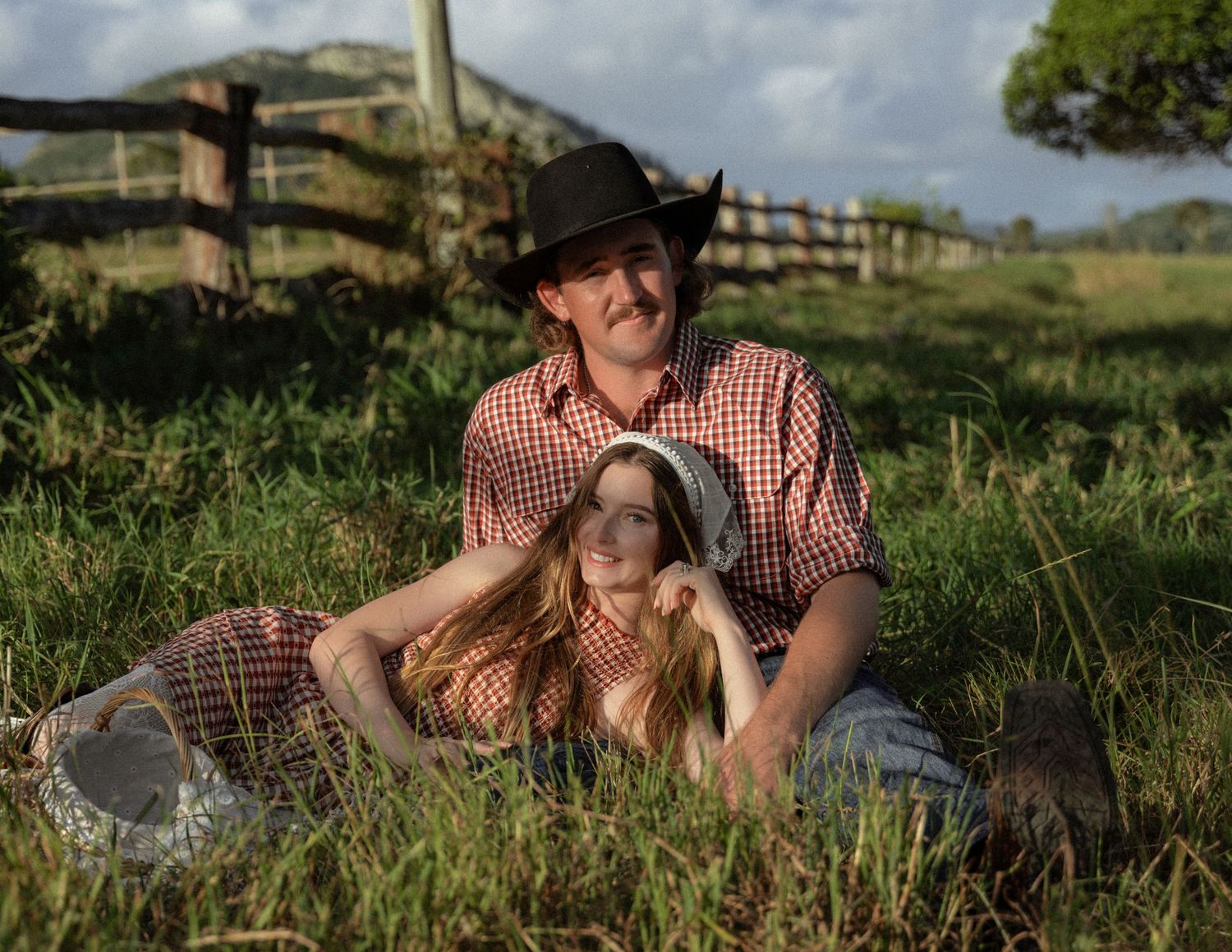 Smiling cowboy in a black hat and plaid shirt sits in a sunny grassy field while a woman in a gingham dress rests against him.