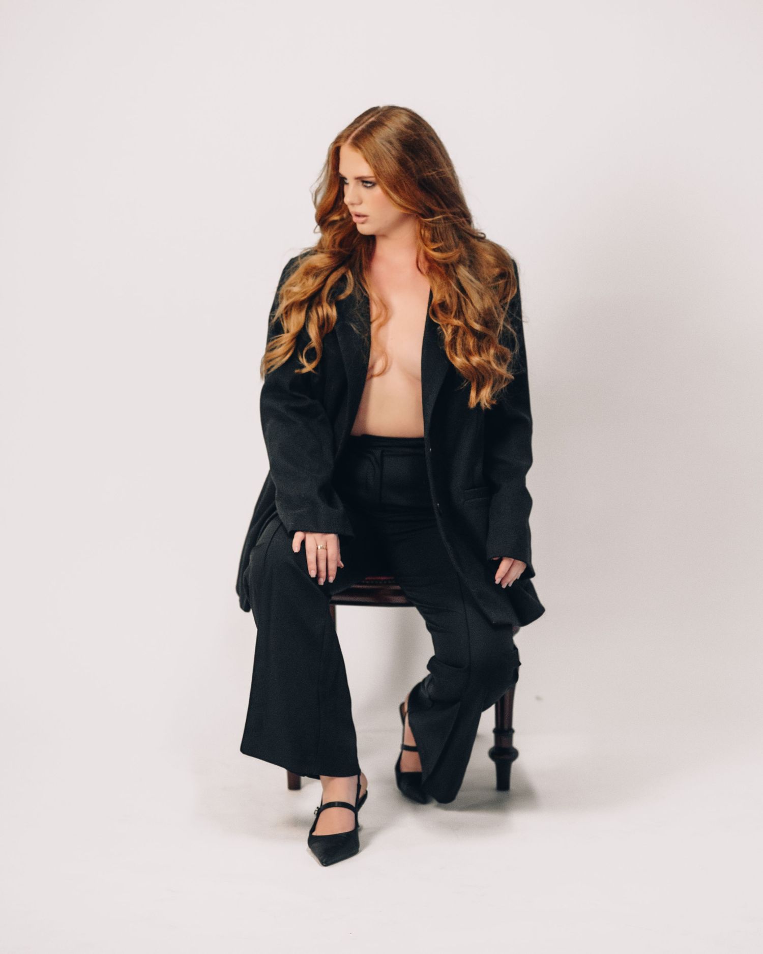 Red-haired woman in an oversized black blazer and trousers sits on a wooden stool against a plain white studio backdrop.