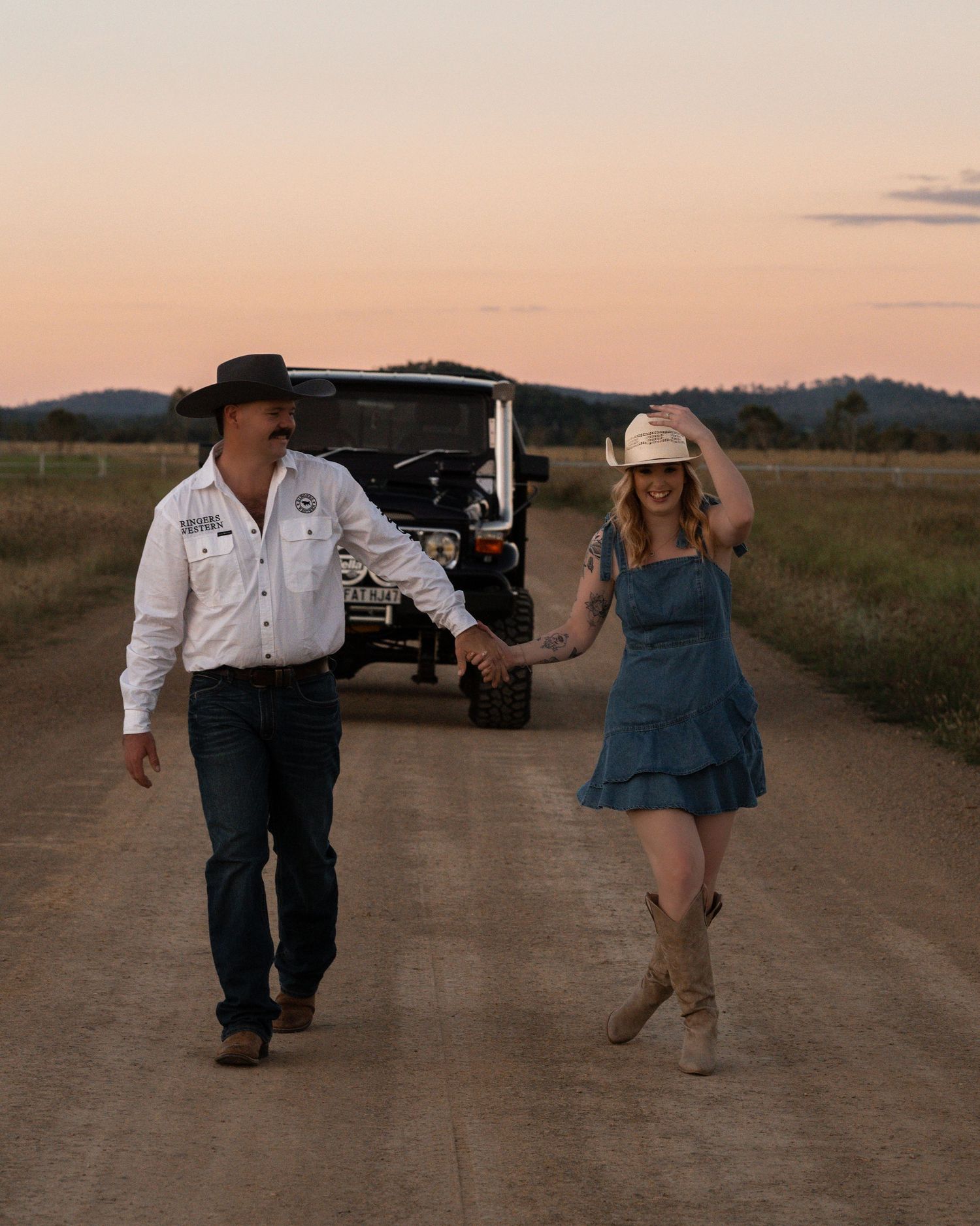 A cowboy and a woman in a denim dress and straw hat hold hands on a country road at sunset beside a 4x4.