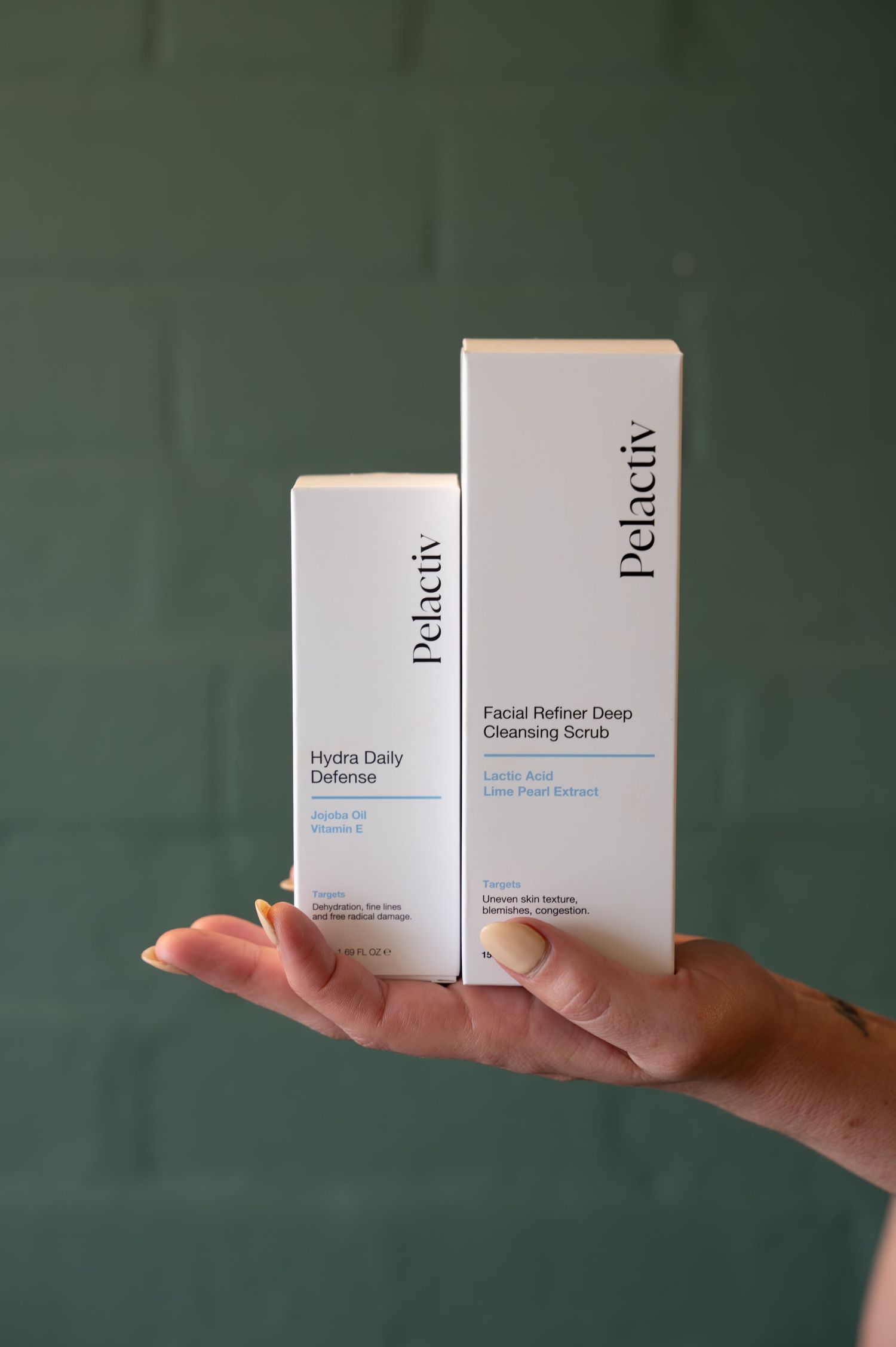 Hand holding two Pelactiv skincare boxes, Hydra Daily Defense and Facial Refiner Deep Cleansing Scrub, against a green brick wall.
