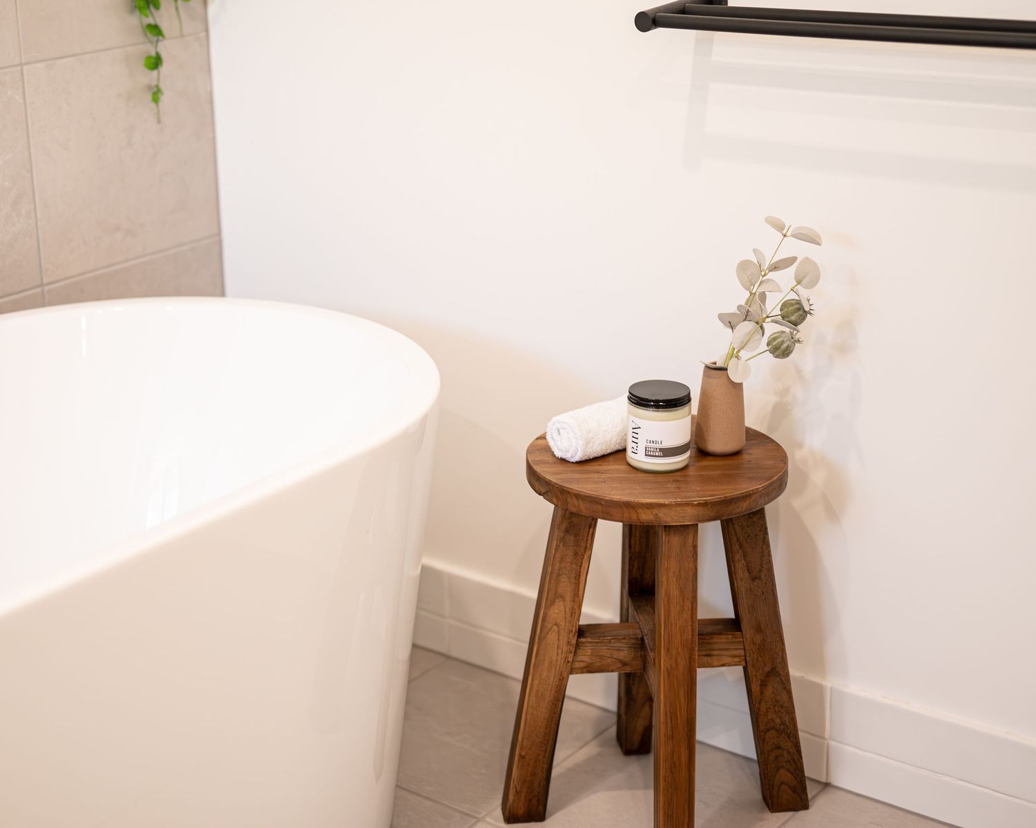 Aura candle and rolled towel on a wooden stool beside a white freestanding bathtub with dried flowers in a vase.