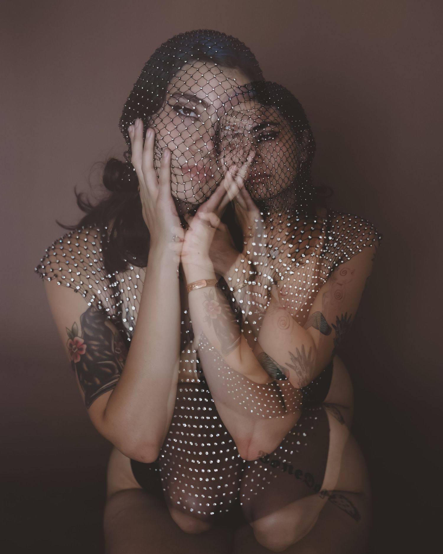 Double-exposure portrait of a tattooed woman in a crystal mesh top and veil, hands framing her face against a brown backdrop.