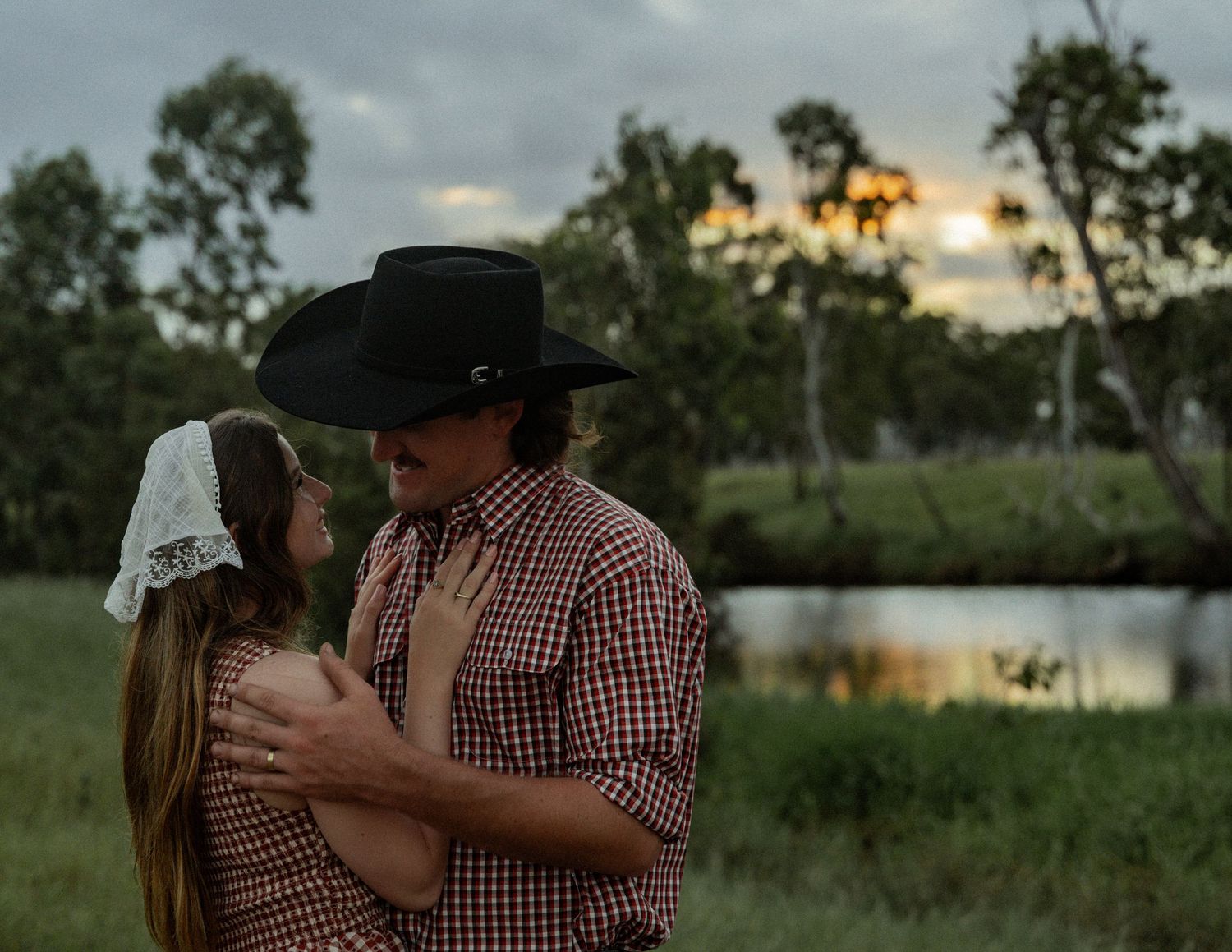 A man in a black cowboy hat and plaid shirt holds a woman in a lace veil near a still pond at sunset.
