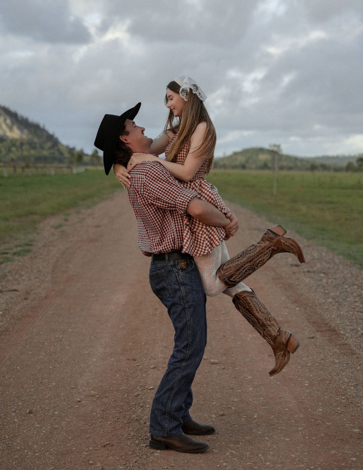 Cowboy in a black hat lifts a laughing woman in a gingham dress and boots on a rural dirt road with green hills behind.