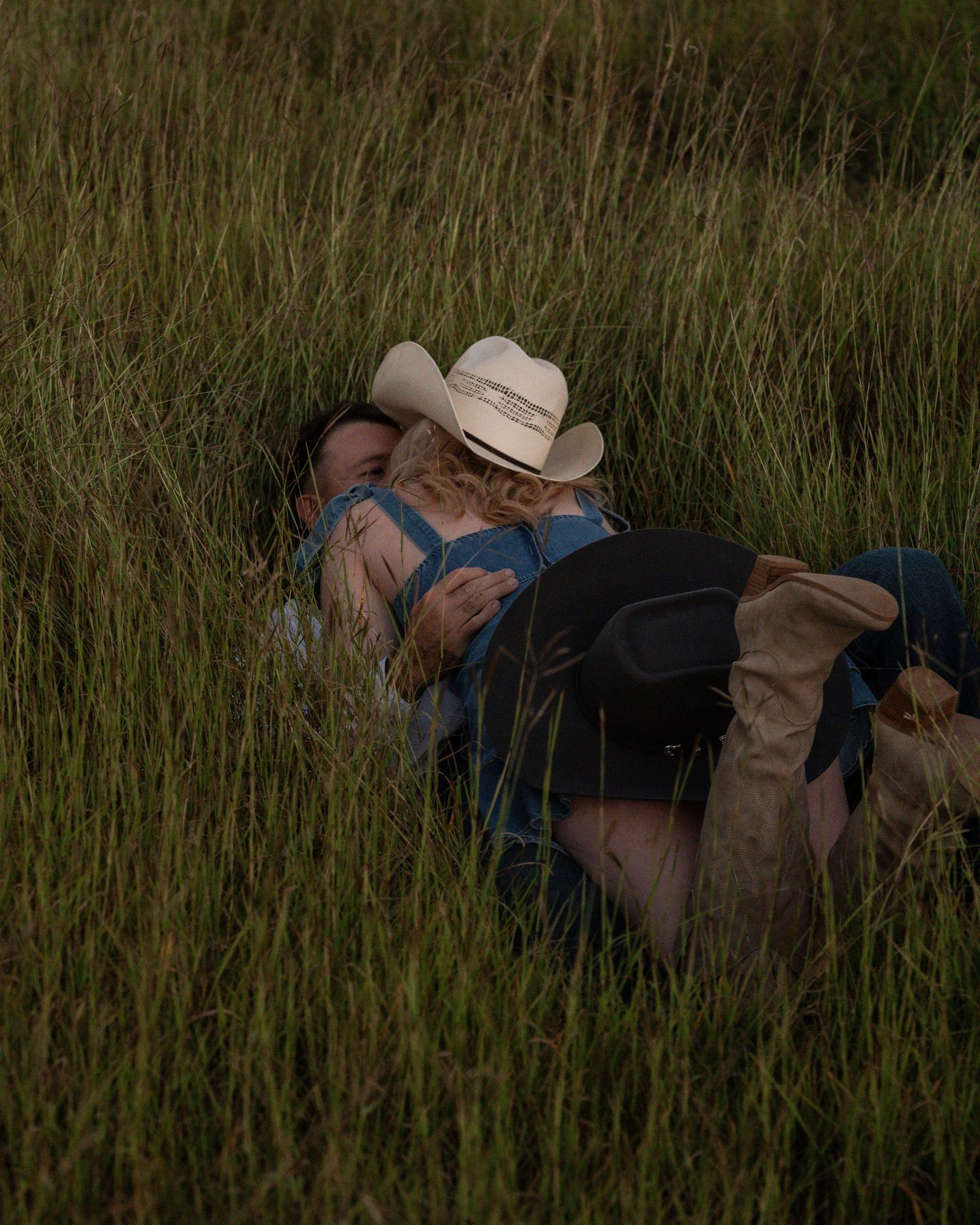 A couple in cowboy hats and boots lie together in tall green grass during a romantic country engagement shoot.