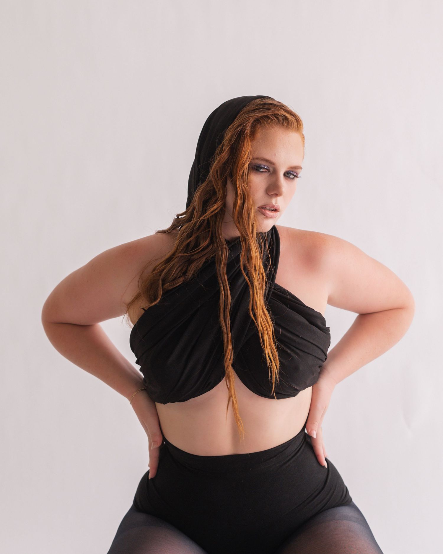 Red-haired woman in a black hooded halter top and tights poses with hands on hips against a soft white studio background.