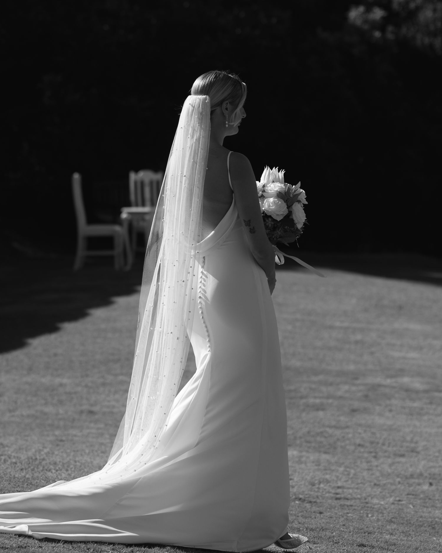 Bride in a elegant white gown and long veil stands facing a garden lawn, holding a bouquet in black and white.