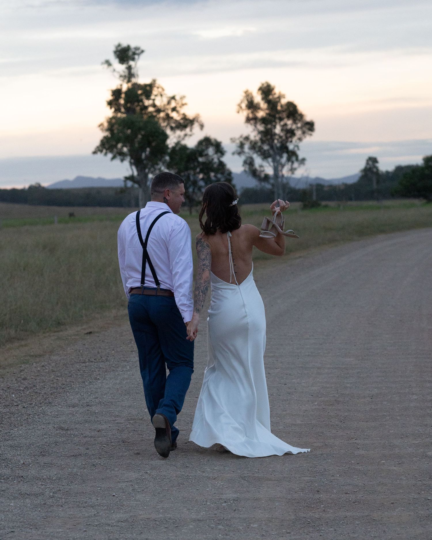 Bride and groom walk together down a quiet gravel road toward distant hills under a soft evening sky.