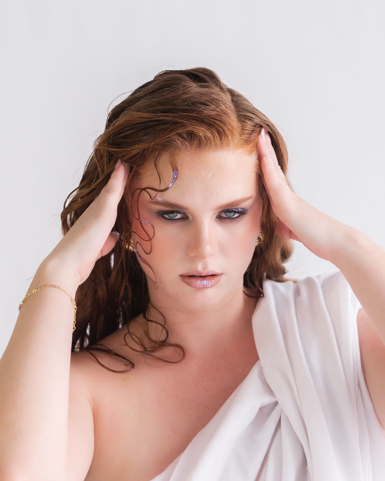 Portrait of a red-haired woman with wet-look hair, bold eye makeup and gold bracelet, hands at her temples, draped in white fabric.