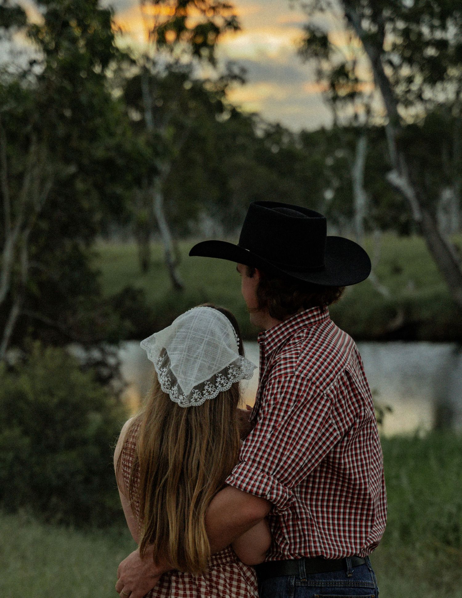 A couple in matching red gingham and cowboy hats embrace while gazing at a calm creek and sunset glow through the trees.