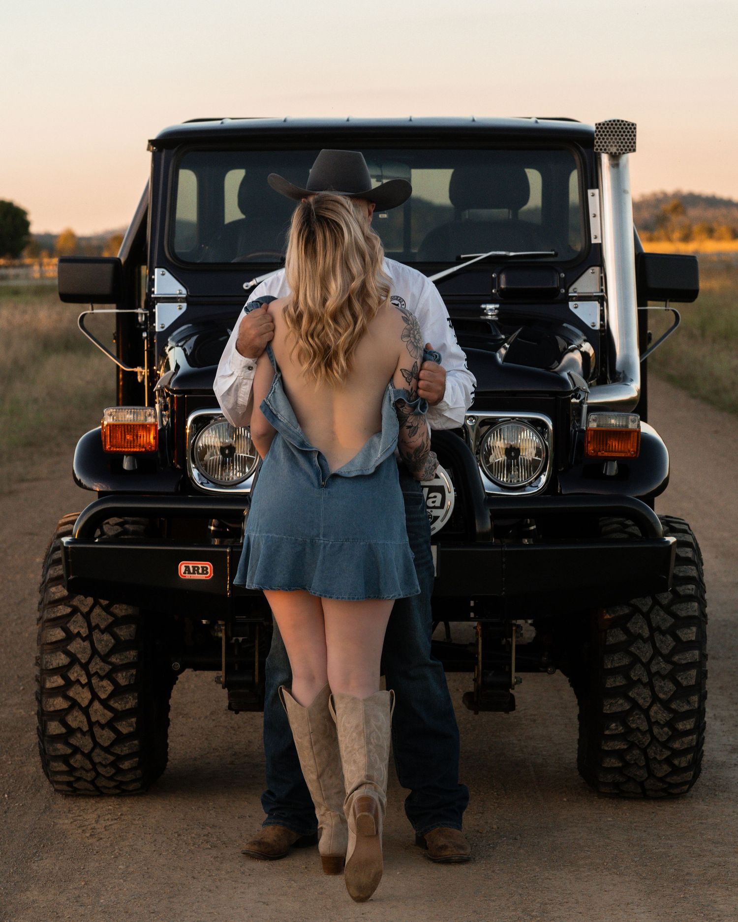 A cowboy in a black hat embraces a woman in a blue dress and boots in front of a black 4x4 on a dirt road.