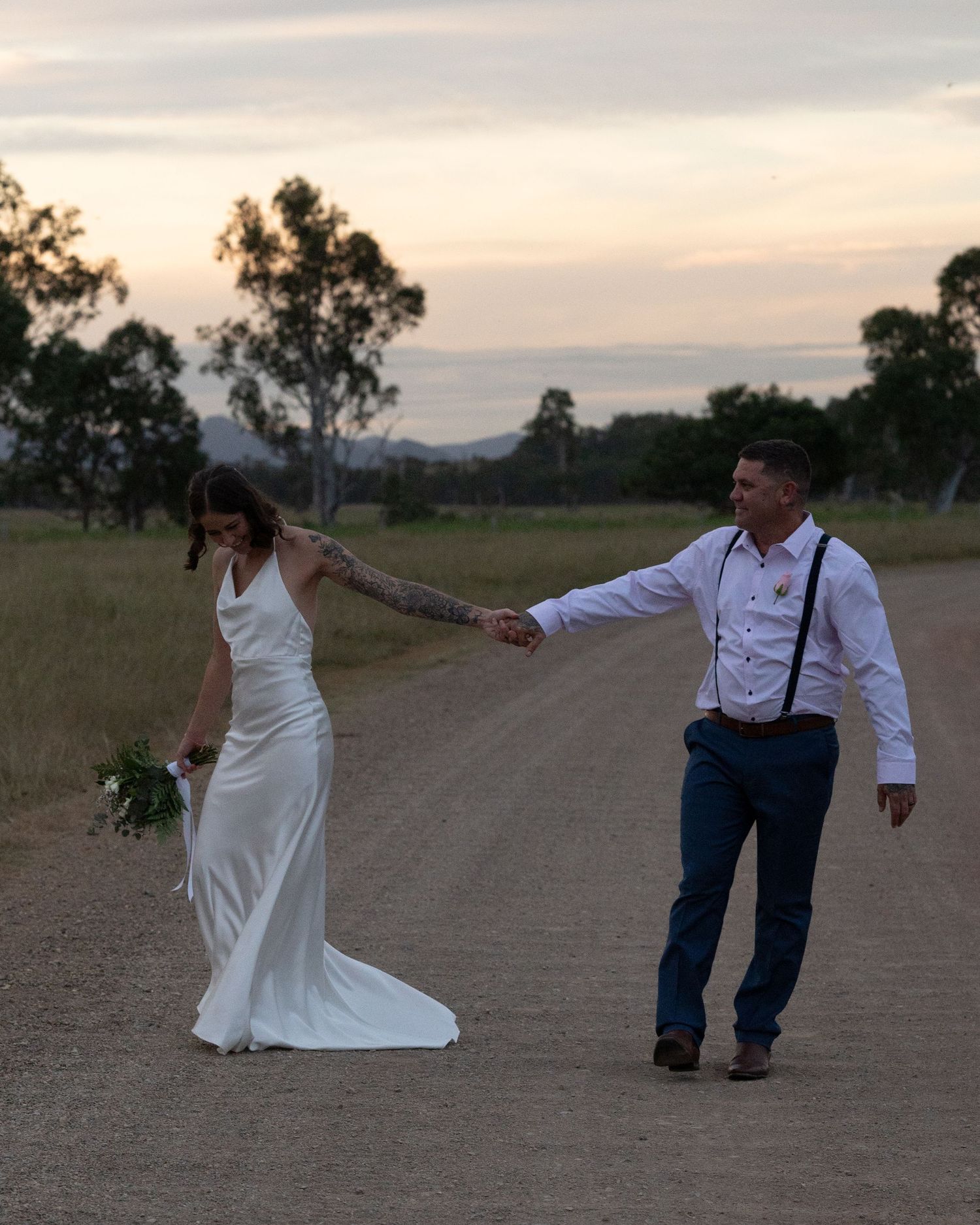 Smiling groom leads his bride, holding a green bouquet, by the hand along a rural gravel road at sunset.