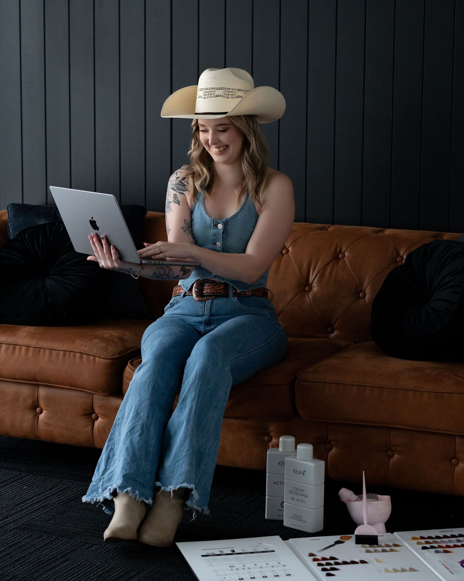 Smiling woman in a cowboy hat and denim outfit sits on a brown leather couch with a laptop, hair color swatches and products on the floor.
