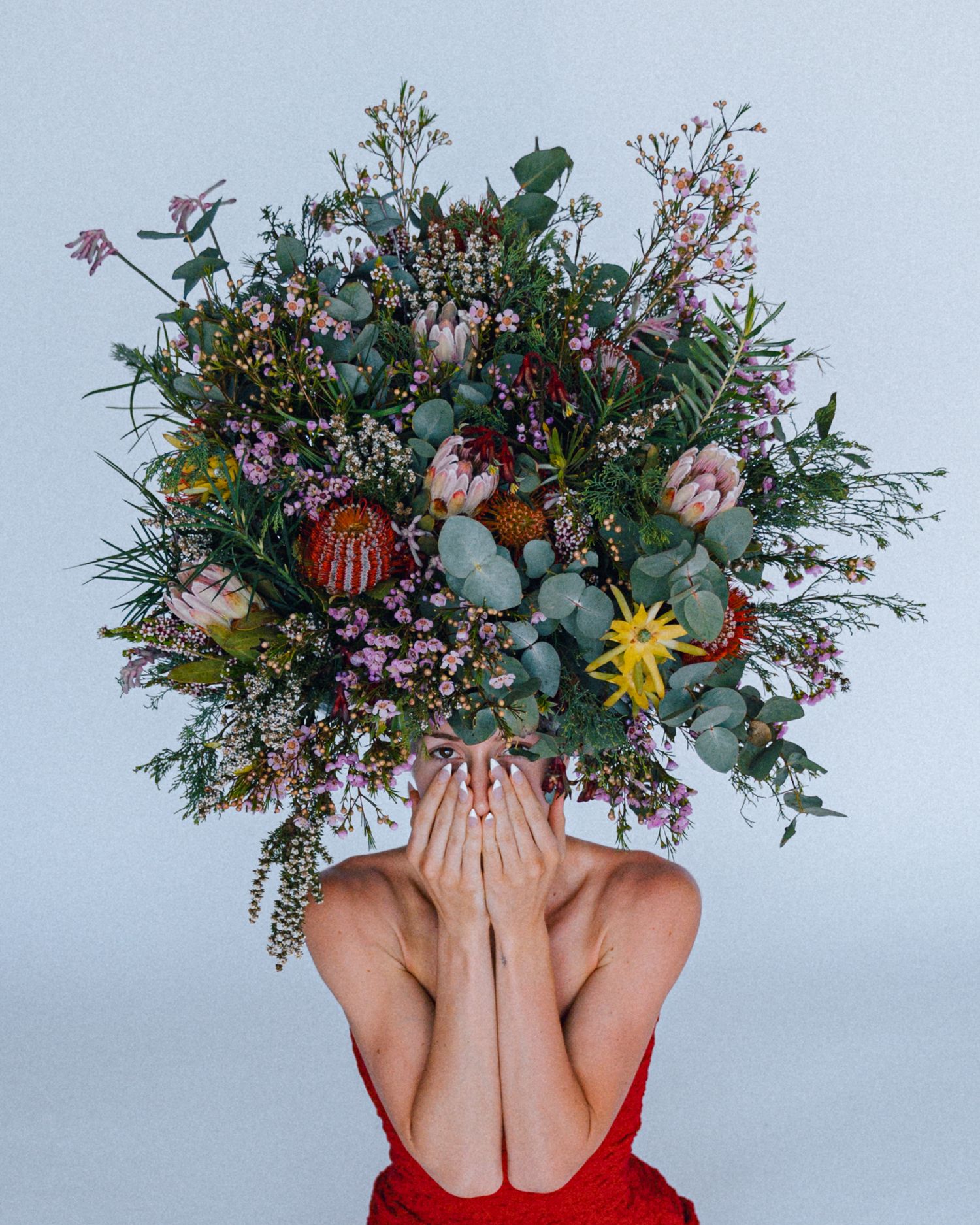 Woman in a red dress covering her face with her hands beneath a large floral arrangement of native flowers and eucalyptus leaves.