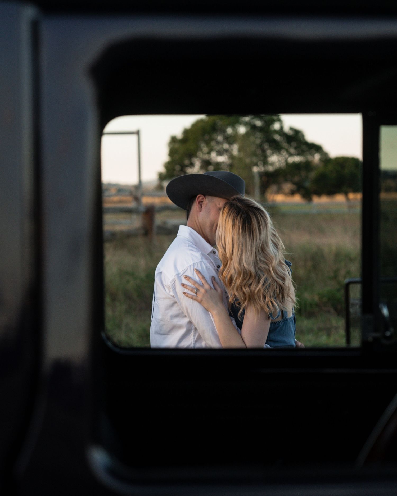 Couple in cowboy attire shares an embrace seen through a vehicle window, with a rural paddock and trees in the background.