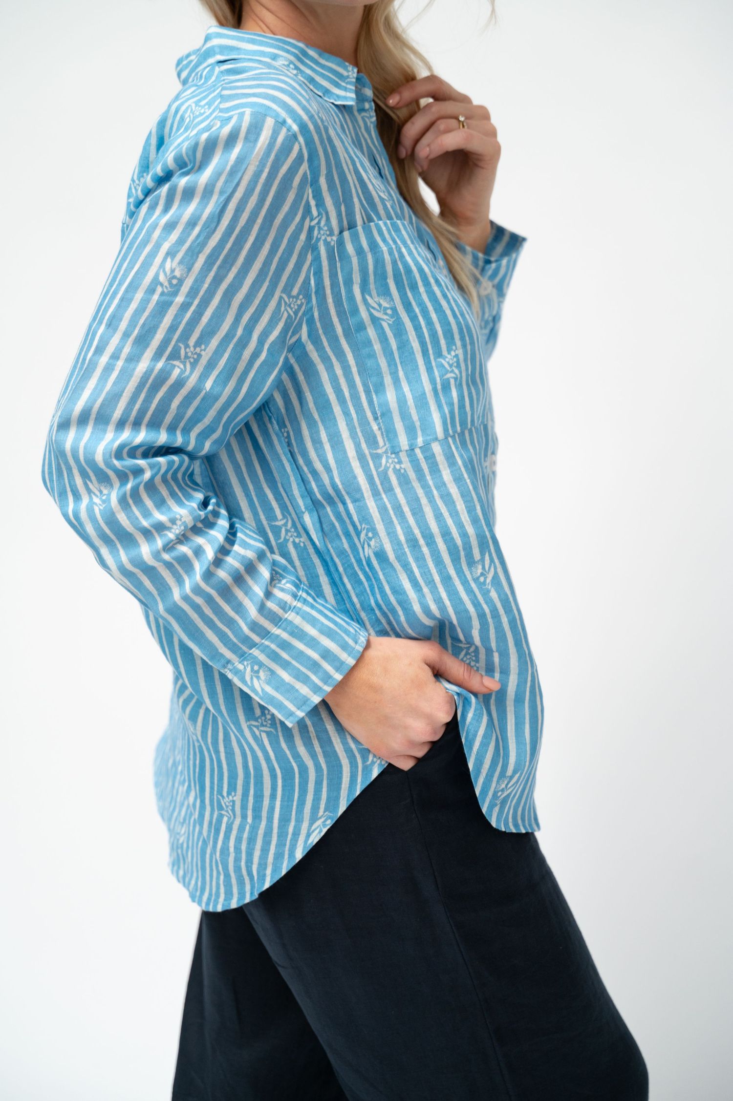 Side view of a woman in a blue and white striped long-sleeve button-up shirt with one hand in the pocket of dark linen pants.