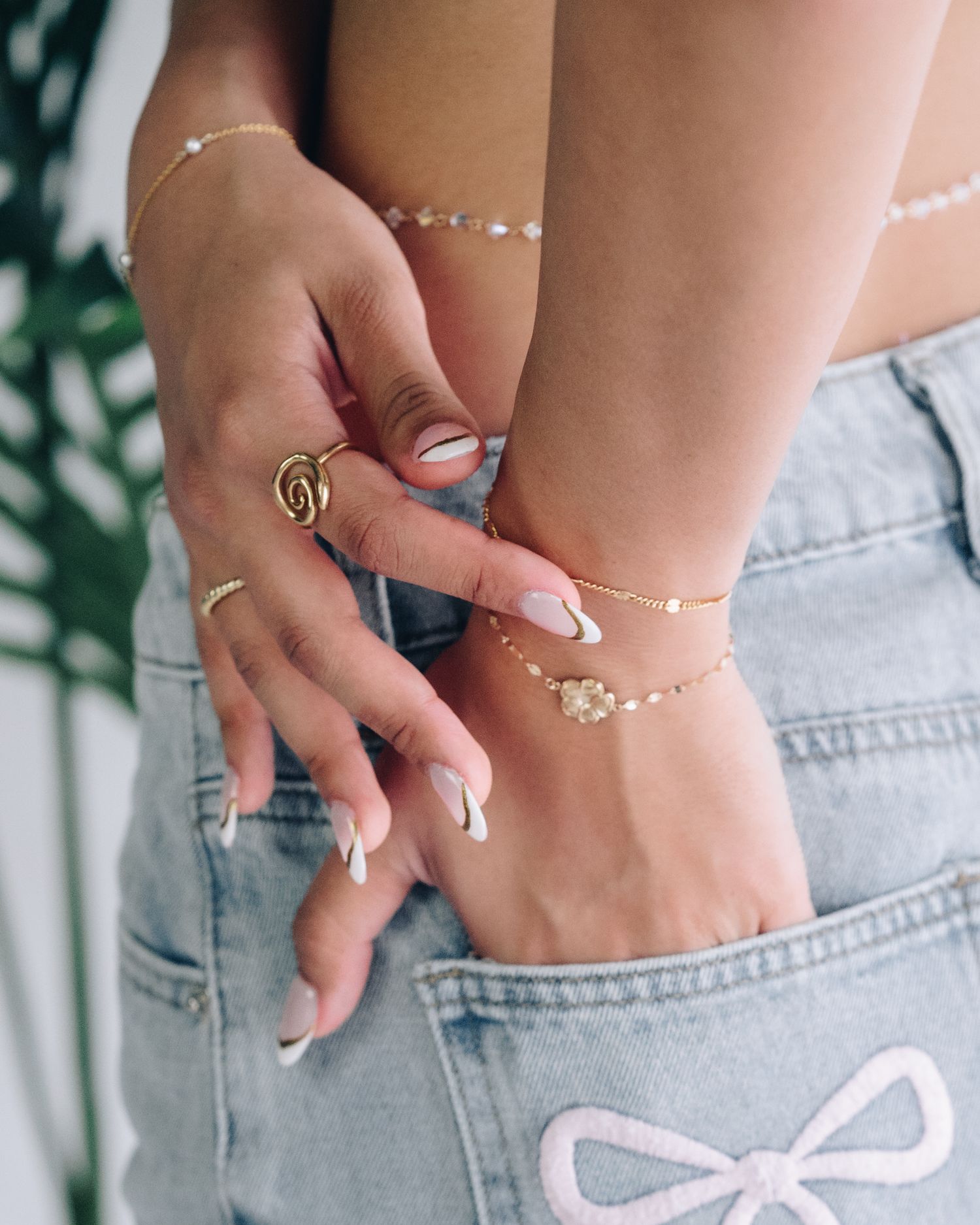 Close-up of a woman's hand in her jeans pocket, wearing gold rings, a delicate gold bracelet and long French-tip nails.