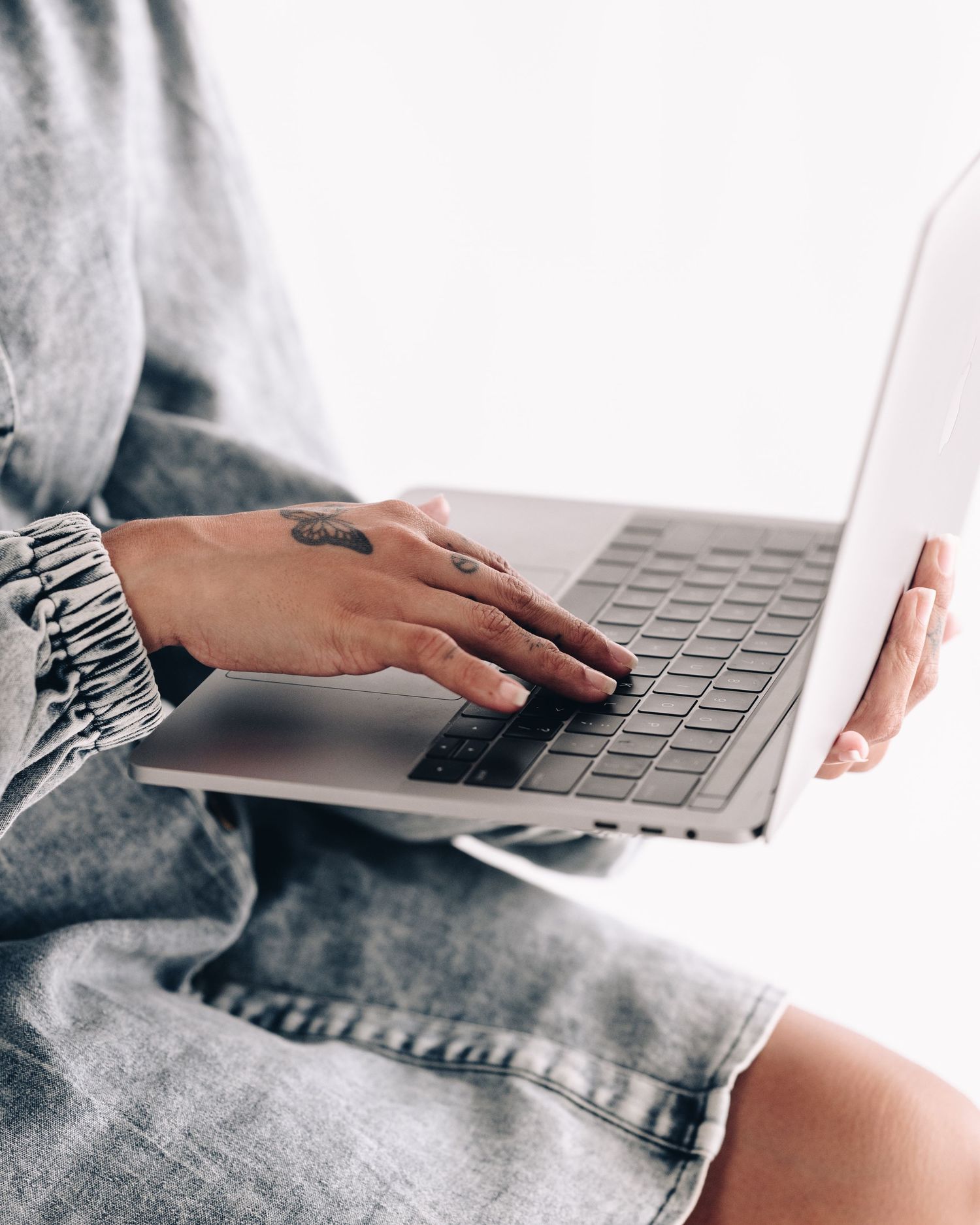 Woman with hand tattoos types on a silver laptop while wearing a gray denim dress.