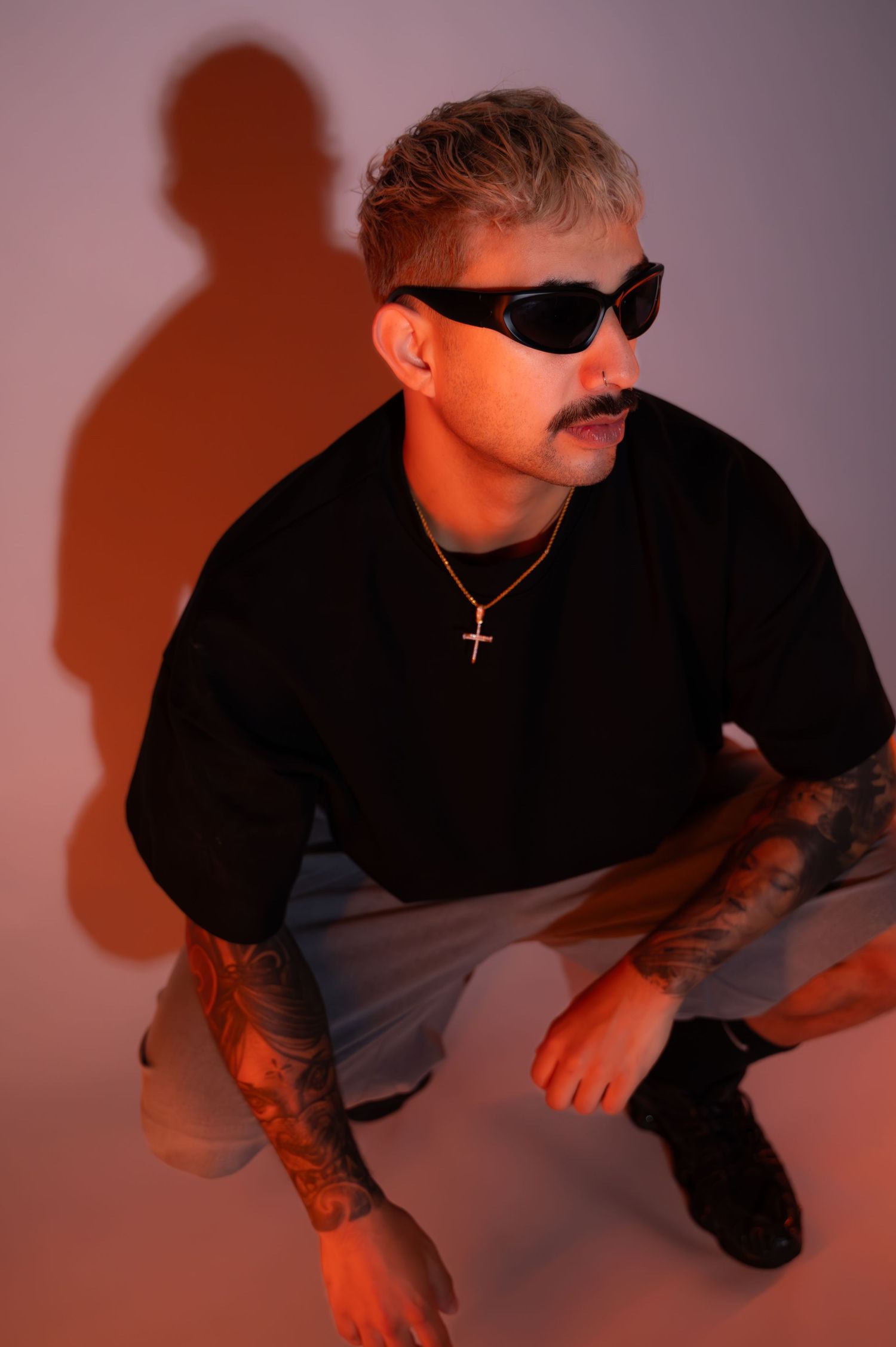 Man with a mustache, wraparound sunglasses and arm tattoos crouches in a black tee and cross necklace against a warm orange studio backdrop.