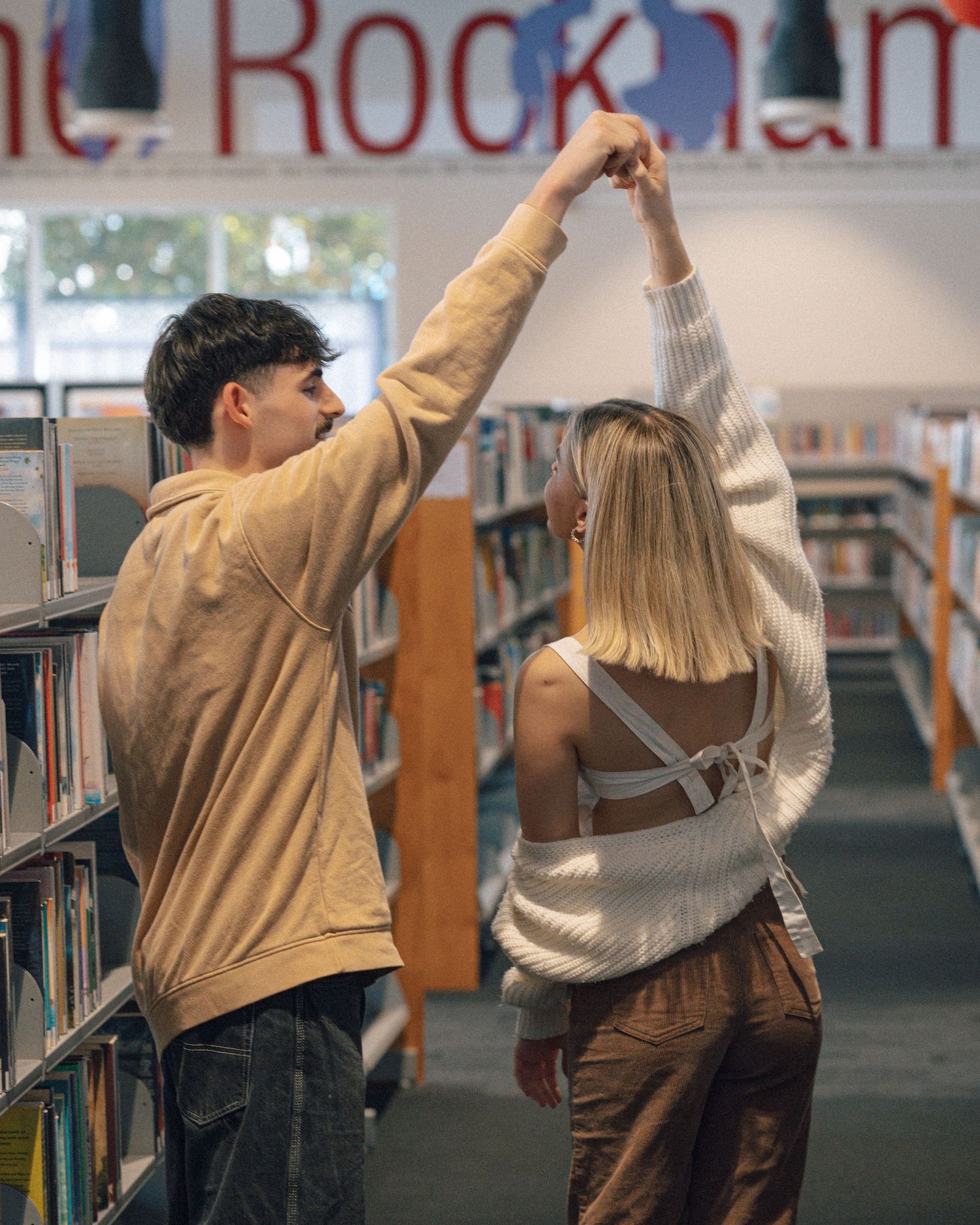 A man in a tan sweatshirt twirls a blonde woman in a white cardigan between library bookshelves.