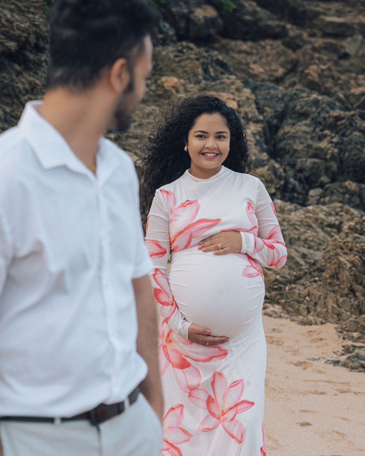 Smiling pregnant woman holds her belly on a rocky beach while her partner in a white shirt stands blurred in the foreground.